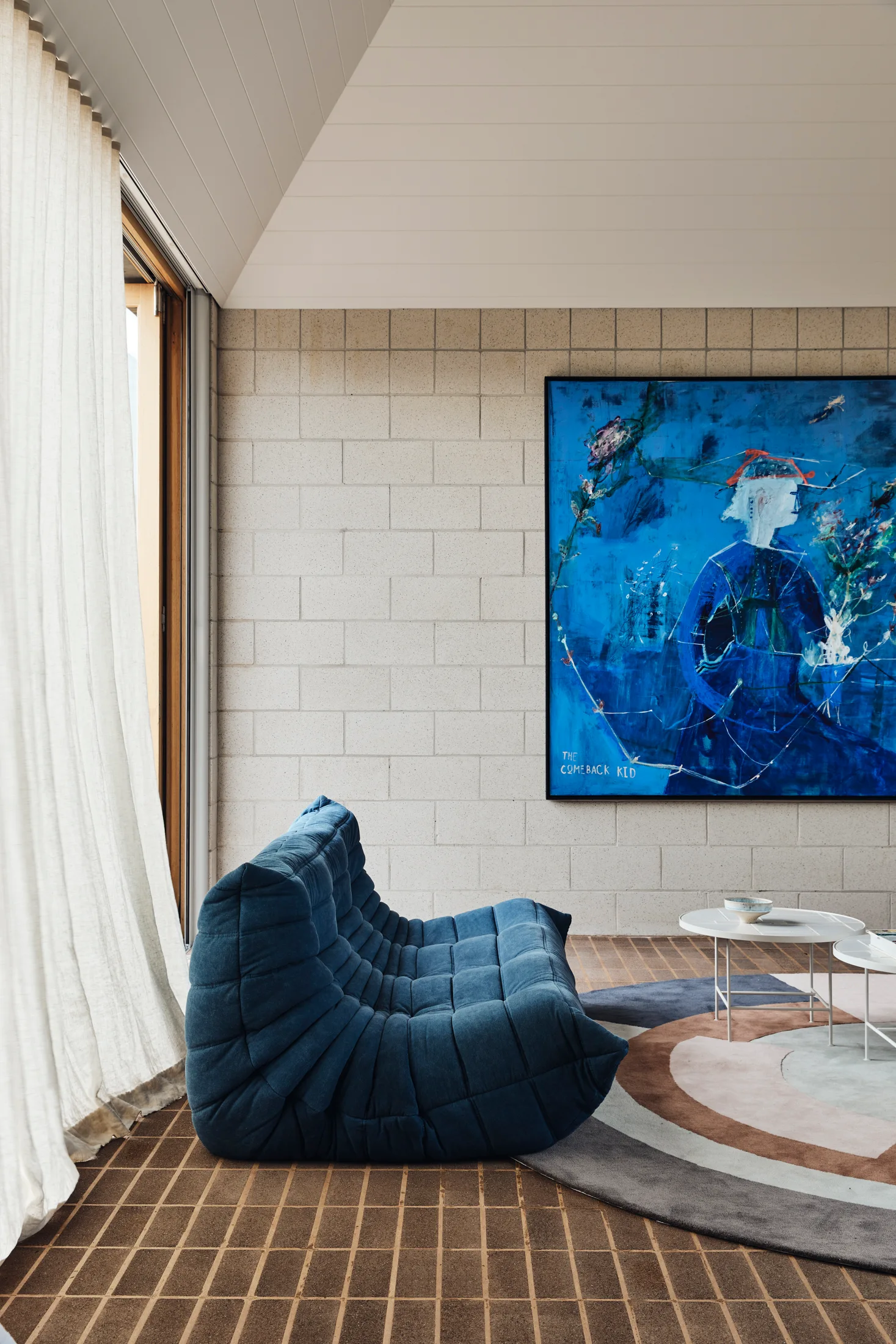 A modern living room features a blue tufted modular sofa set against a light grey masonry block wall adorned with a large blue abstract painting.