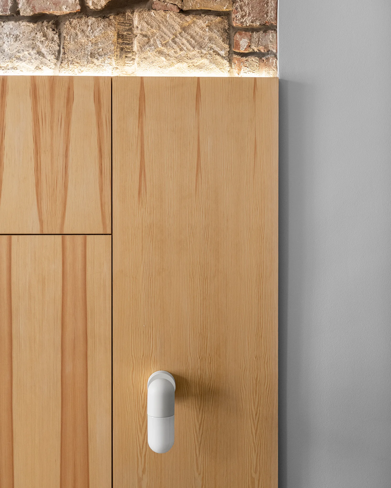 A close-up of vertical grain wood cabinets featuring a white cylindrical handle, positioned under an exposed brick wall highlighted by LED strip lighting.