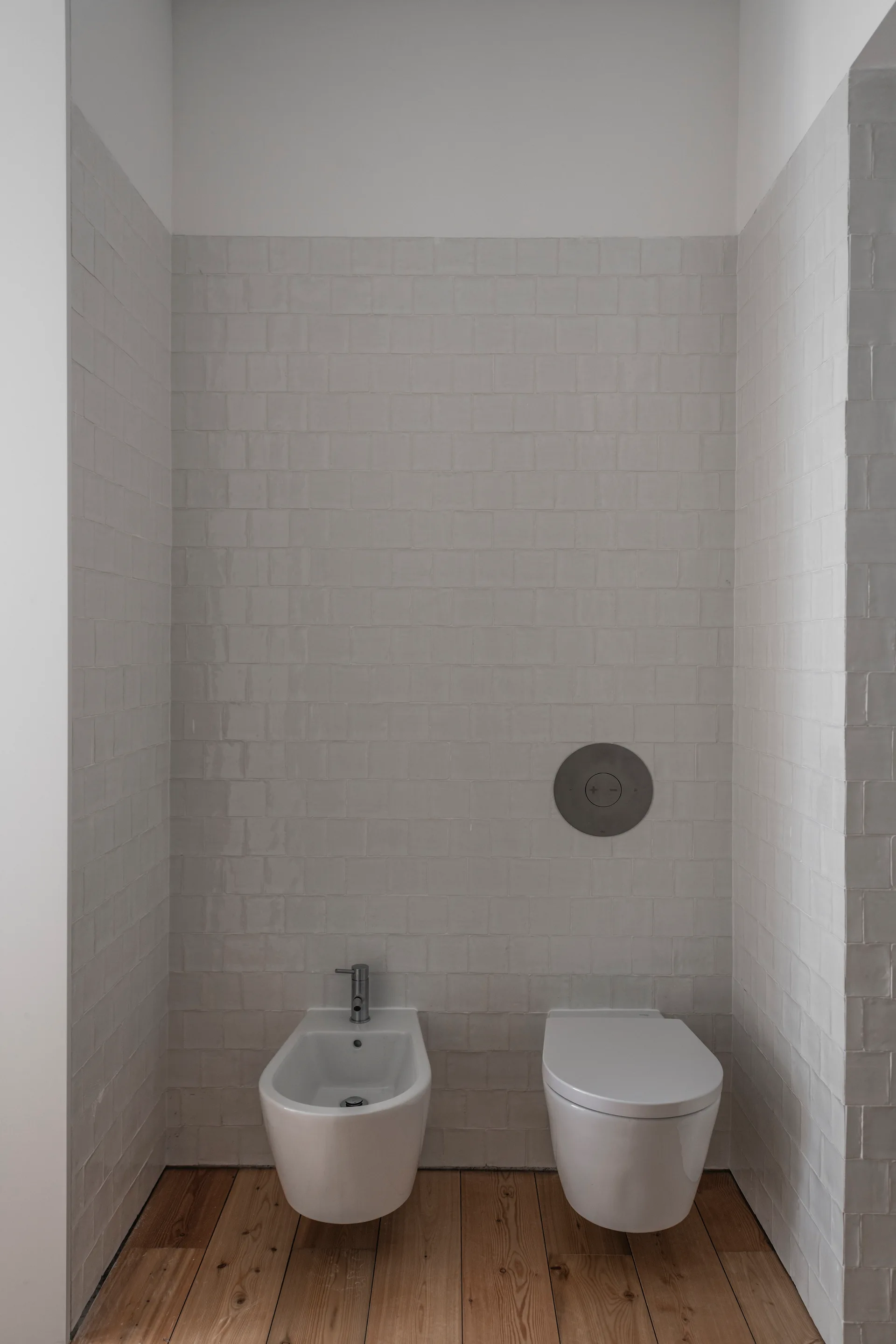 A minimalist bathroom alcove features a white wall-mounted toilet and bidet against glossy square tiled walls with natural wood plank flooring.