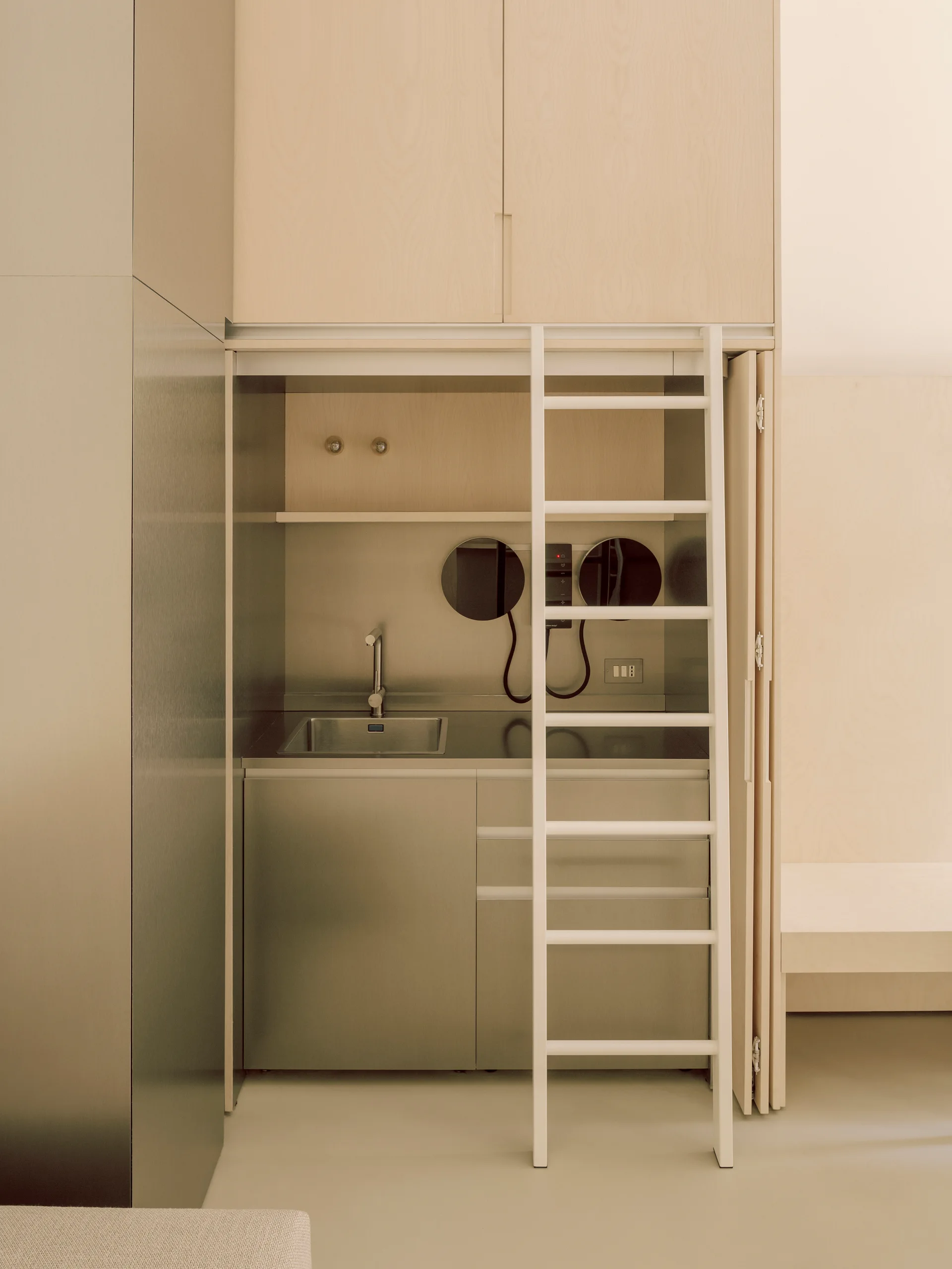 A compact kitchenette features a stainless steel sink, light wood upper cabinetry, a metal backsplash with circular mirrors, and a white ladder.