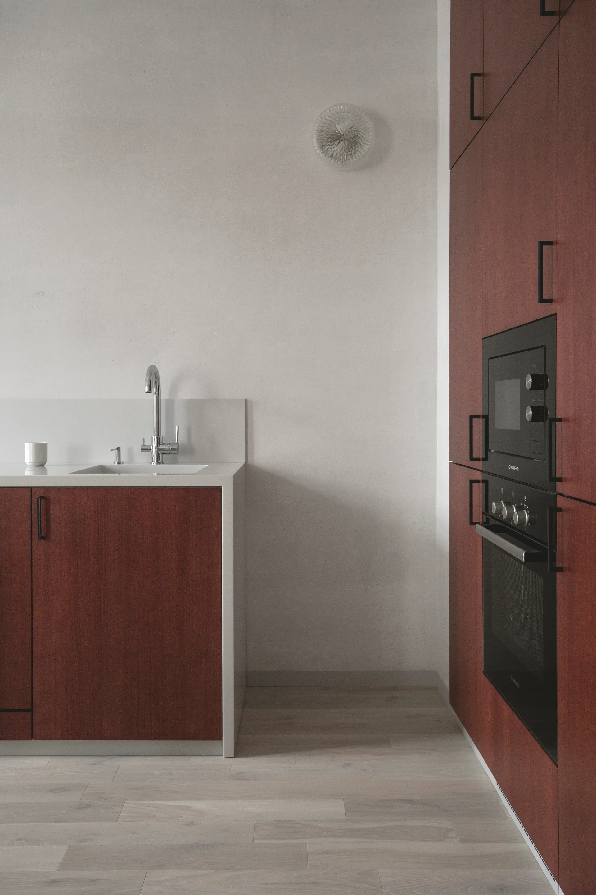 A minimalist kitchen features dark red wood cabinetry, an integrated sink with a chrome faucet, light wood flooring, and a textured white wall.