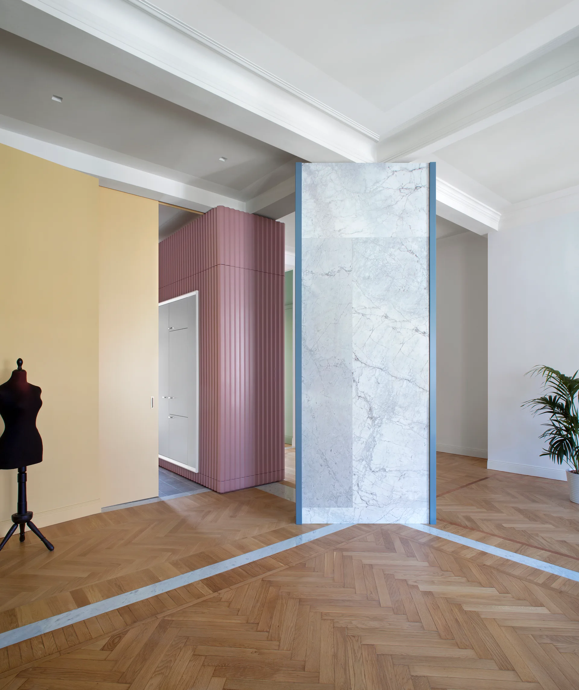 A spacious room features a freestanding marble partition, a dusty pink fluted wall, a yellow panel, and herringbone wood flooring.