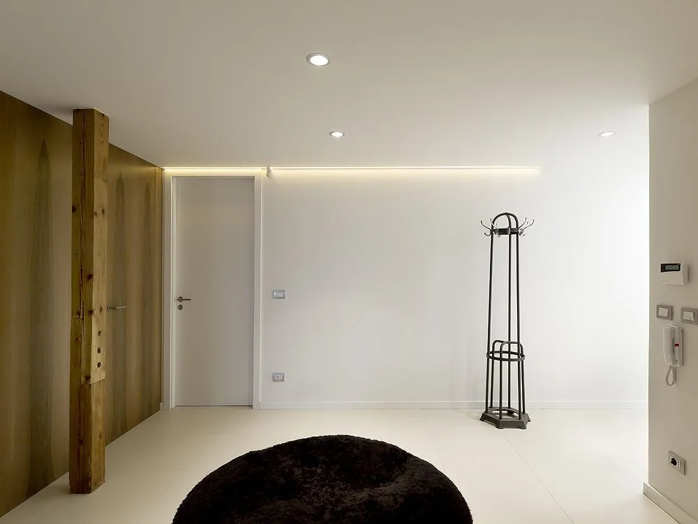 A minimalist entryway featuring a rustic vertical wood post, a dark metal coat rack, white walls with a hidden light strip, and large white floor tiles.