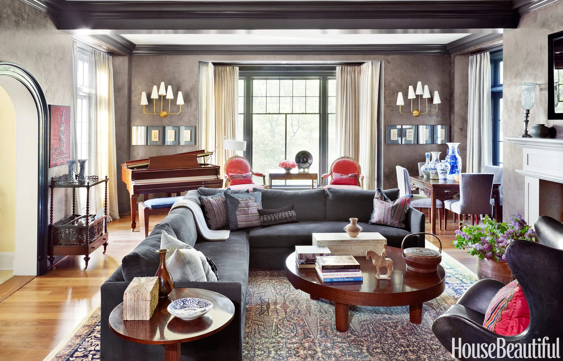 A spacious living room features a charcoal sectional sofa, a round wooden coffee table, textured grey walls, and a grand piano near the window.