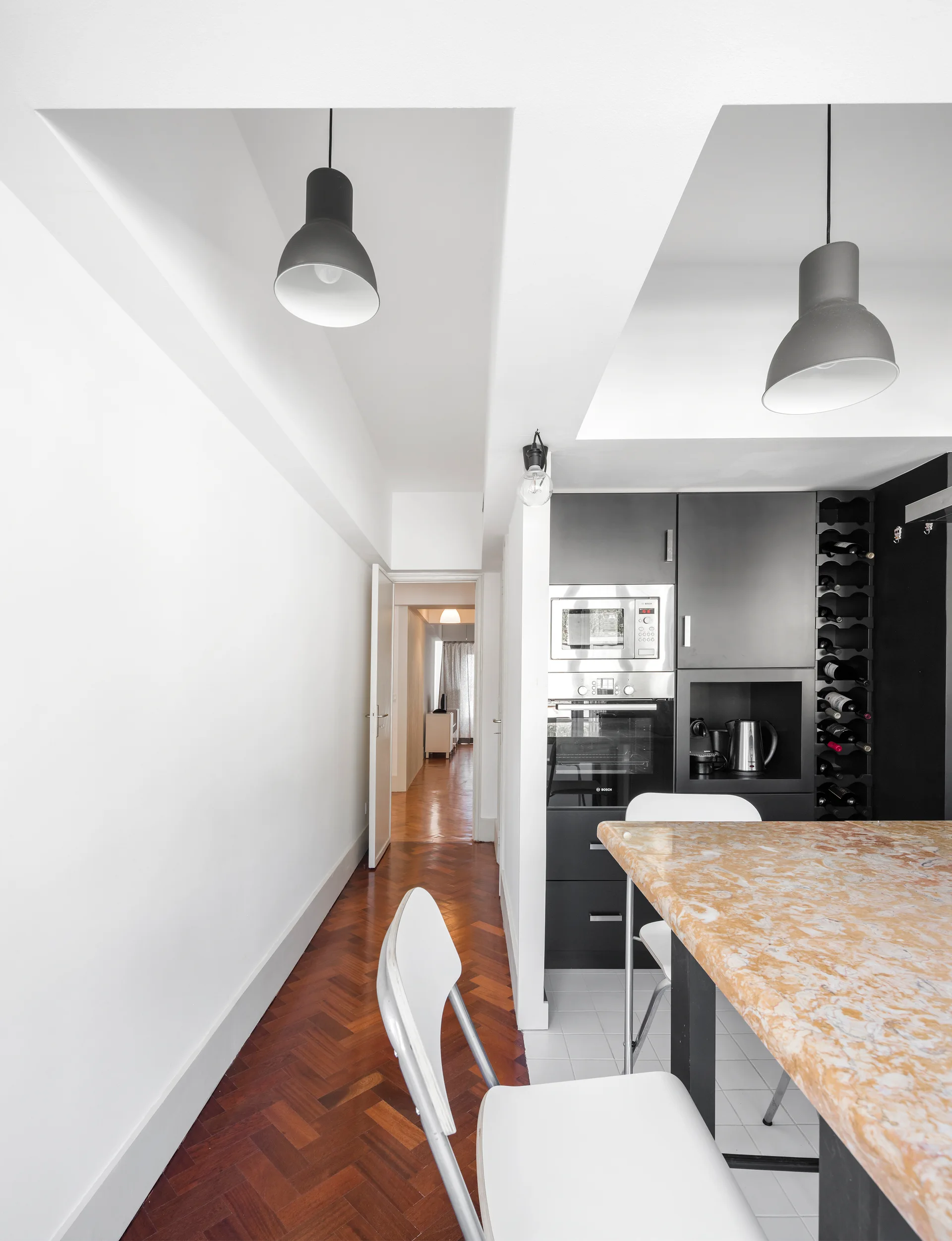 A modern kitchen featuring a marble-patterned island countertop and dark cabinets next to a hallway with herringbone parquet floors.