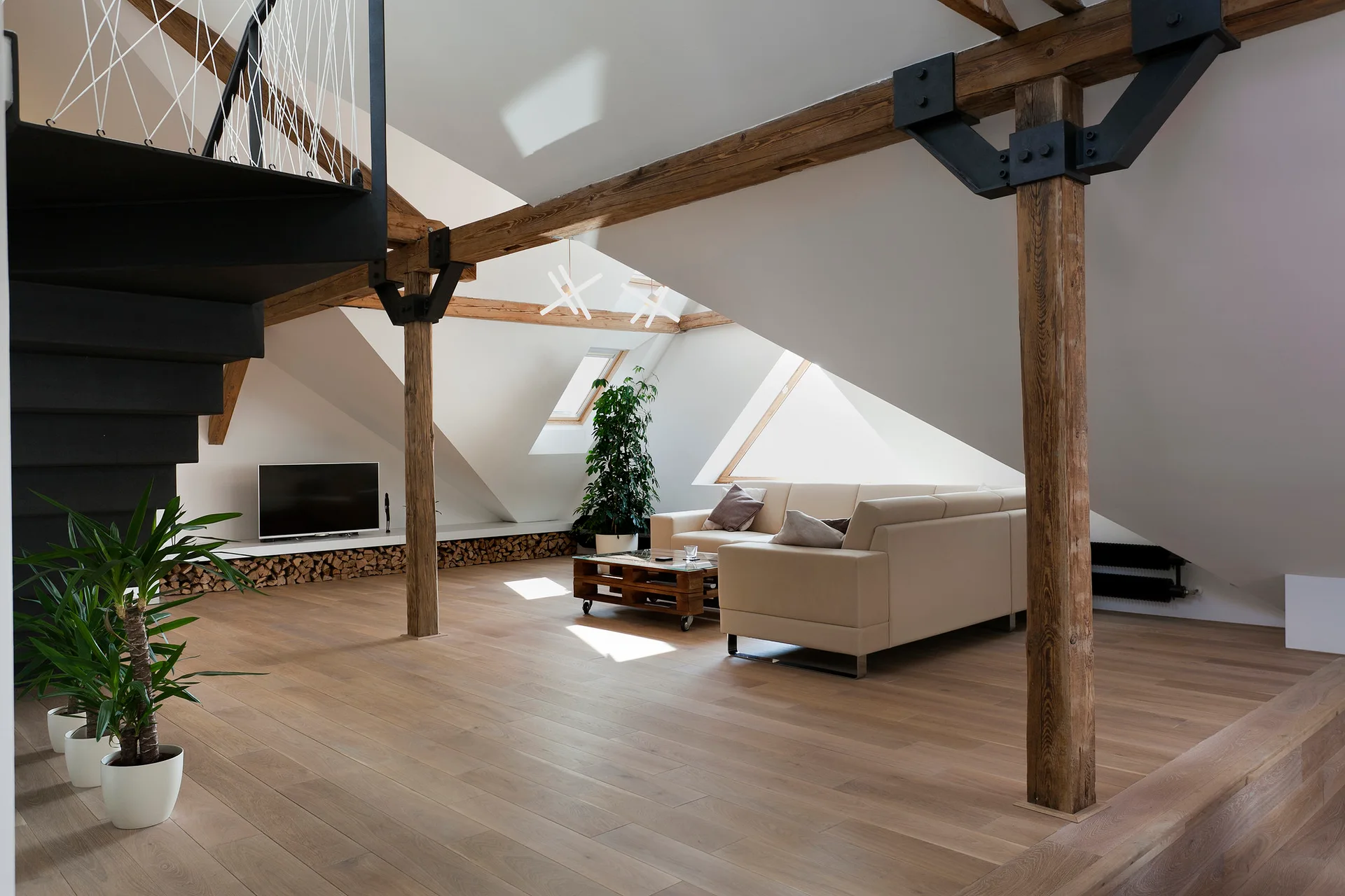 A spacious attic living room featuring a beige leather sectional sofa, exposed structural timber beams, and a black steel spiral staircase.