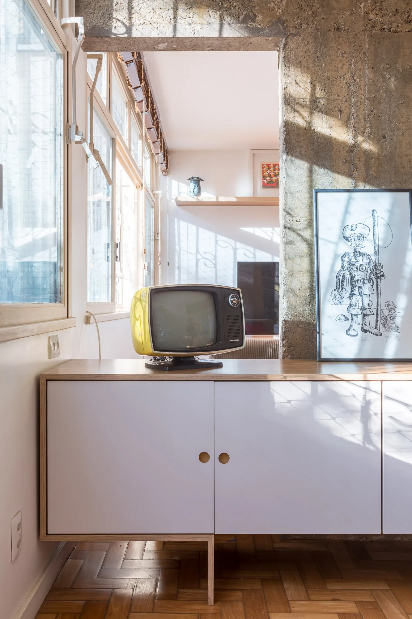 A vintage yellow television sits on a white and light-wood sideboard next to a framed line-art drawing and a rough exposed concrete pillar.