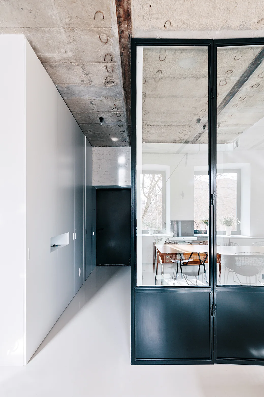 A modern hallway featuring seamless white built-in closets, an exposed concrete ceiling, and a black metal glass partition looking into a home office.
