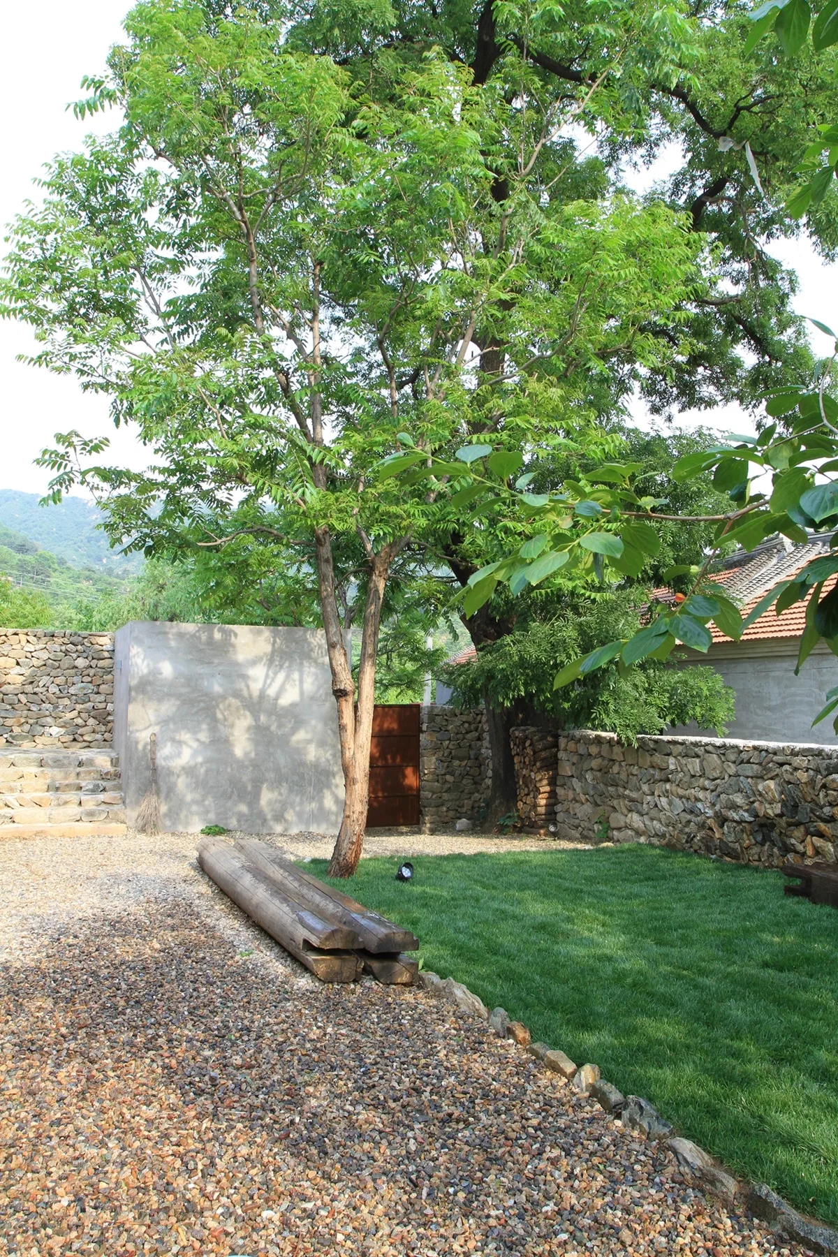 A rustic garden features a green lawn, gravel ground cover, a large tree, fieldstone walls, and a smooth concrete accent wall.