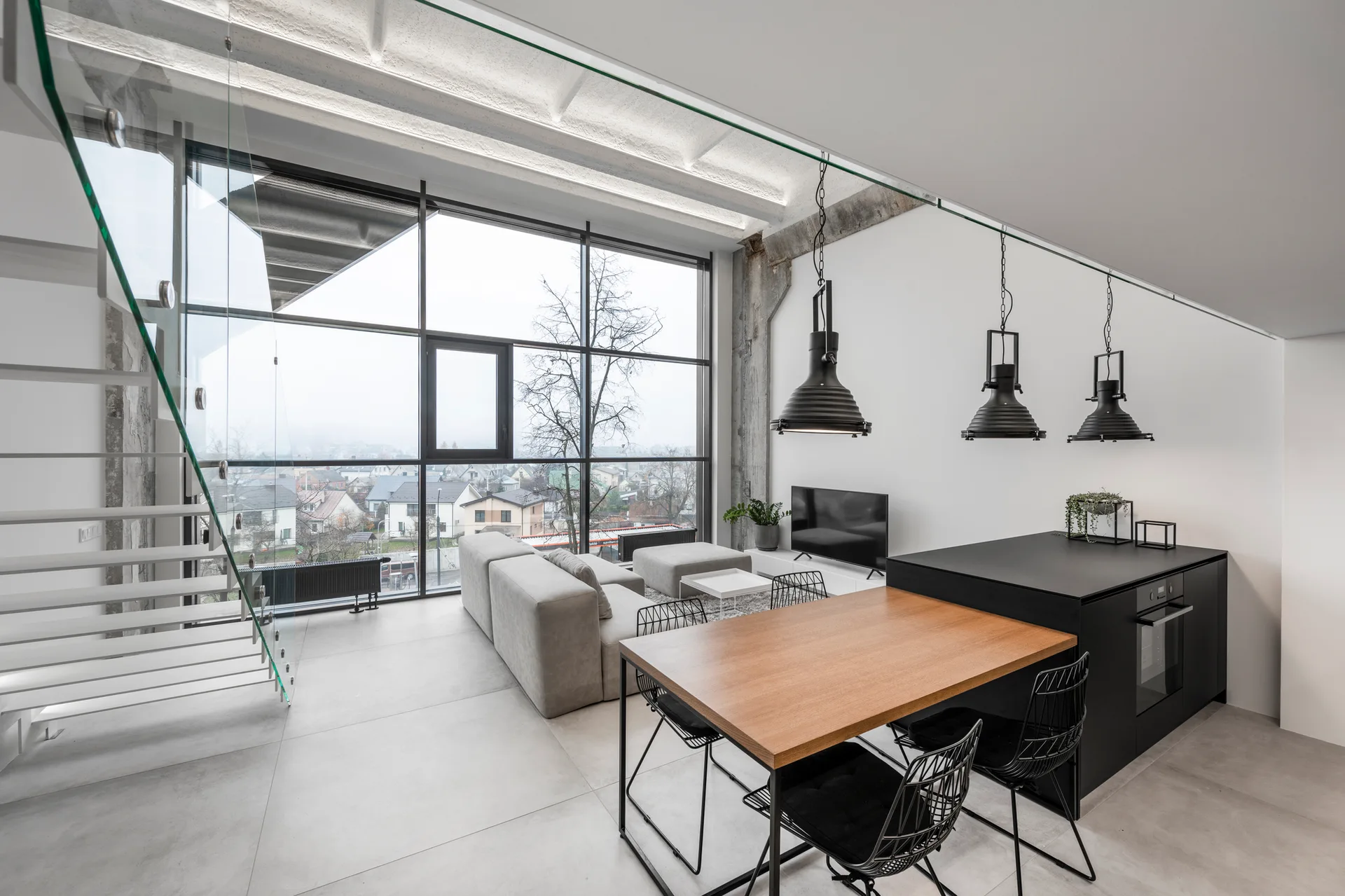 Modern open-concept room featuring a glass-railing staircase, a gray sectional sofa, and a black kitchen island with an attached wood dining table.