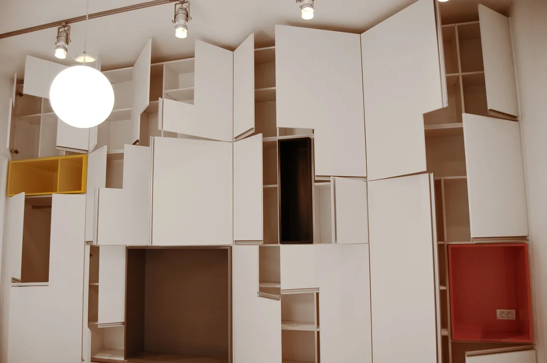 A large white modular wall cabinet system featuring asymmetric doors, yellow and red accent cubbies, and a hanging globe pendant light.