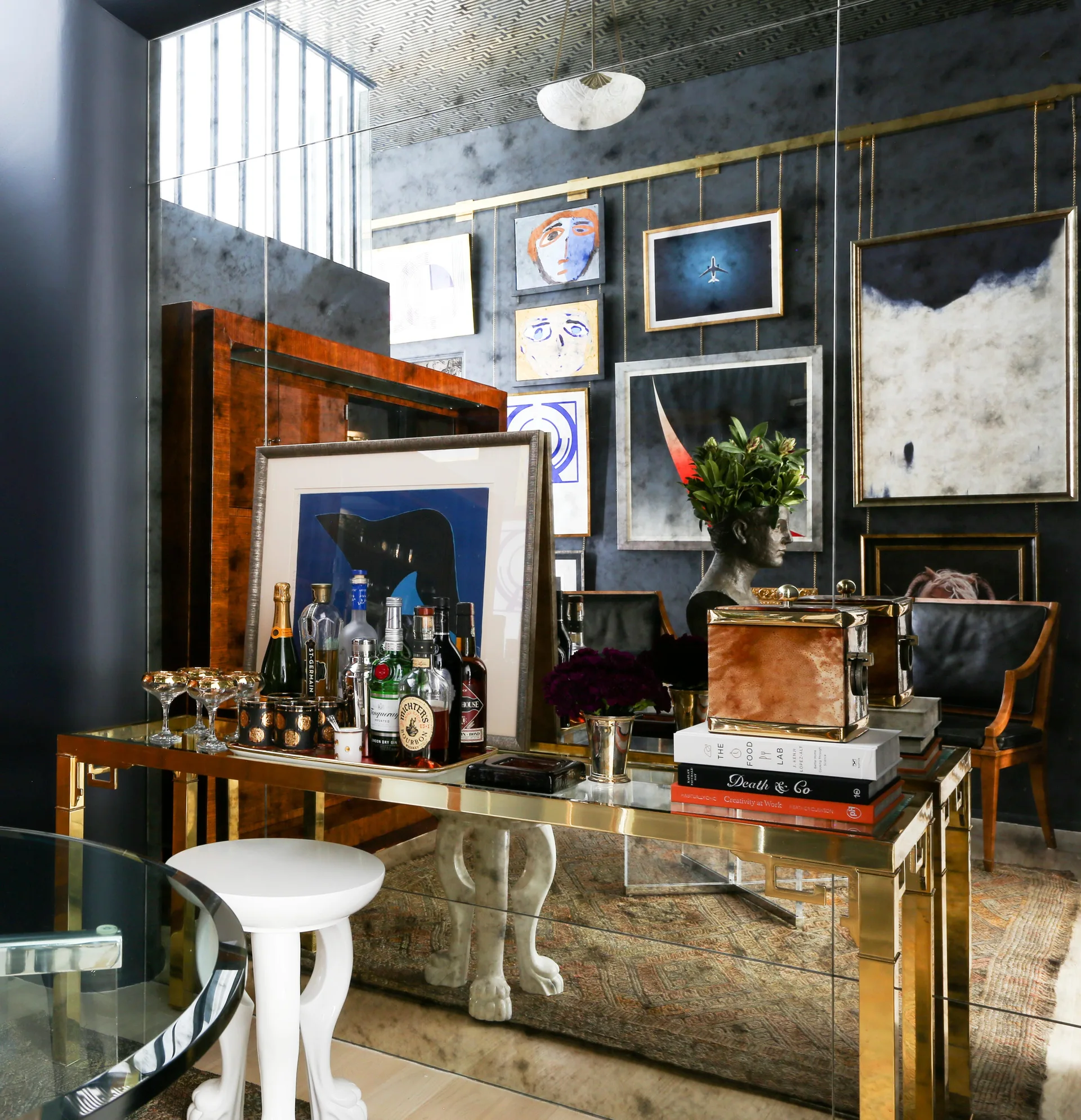 A gold console table styled with liquor bottles and books sits in front of a dark mirrored gallery wall hung with eclectic framed artwork.