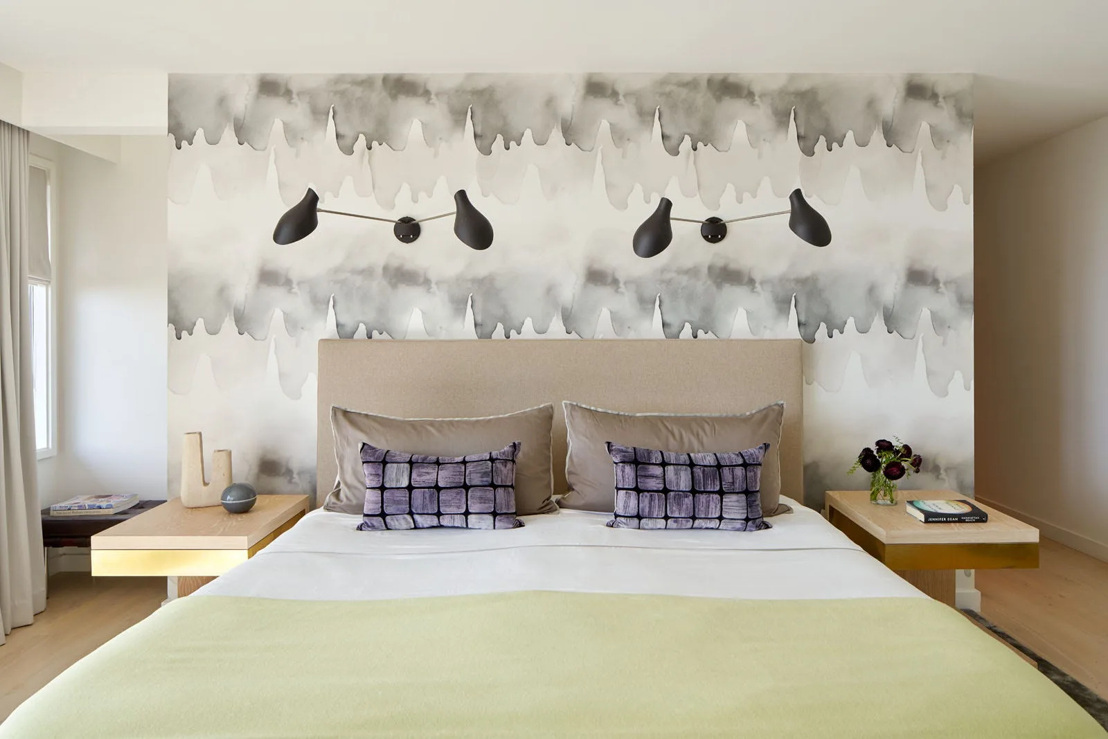 A modern bedroom features a bed with a beige headboard, abstract watercolor wallpaper, two black wall sconces, and floating wood nightstands.
