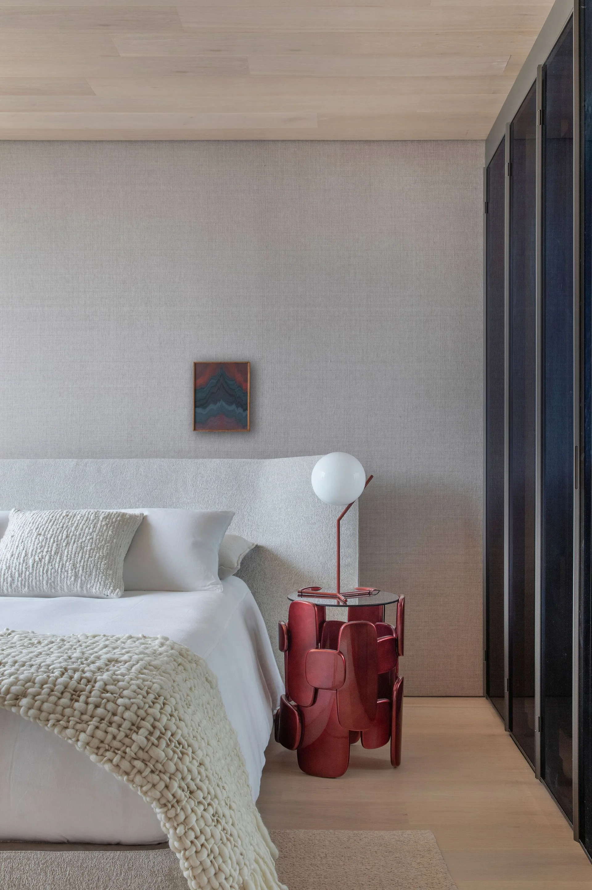 A minimalist bedroom featuring a textured off-white bed, a small framed art piece on a beige wallpapered wall, and a sculptural red side table.