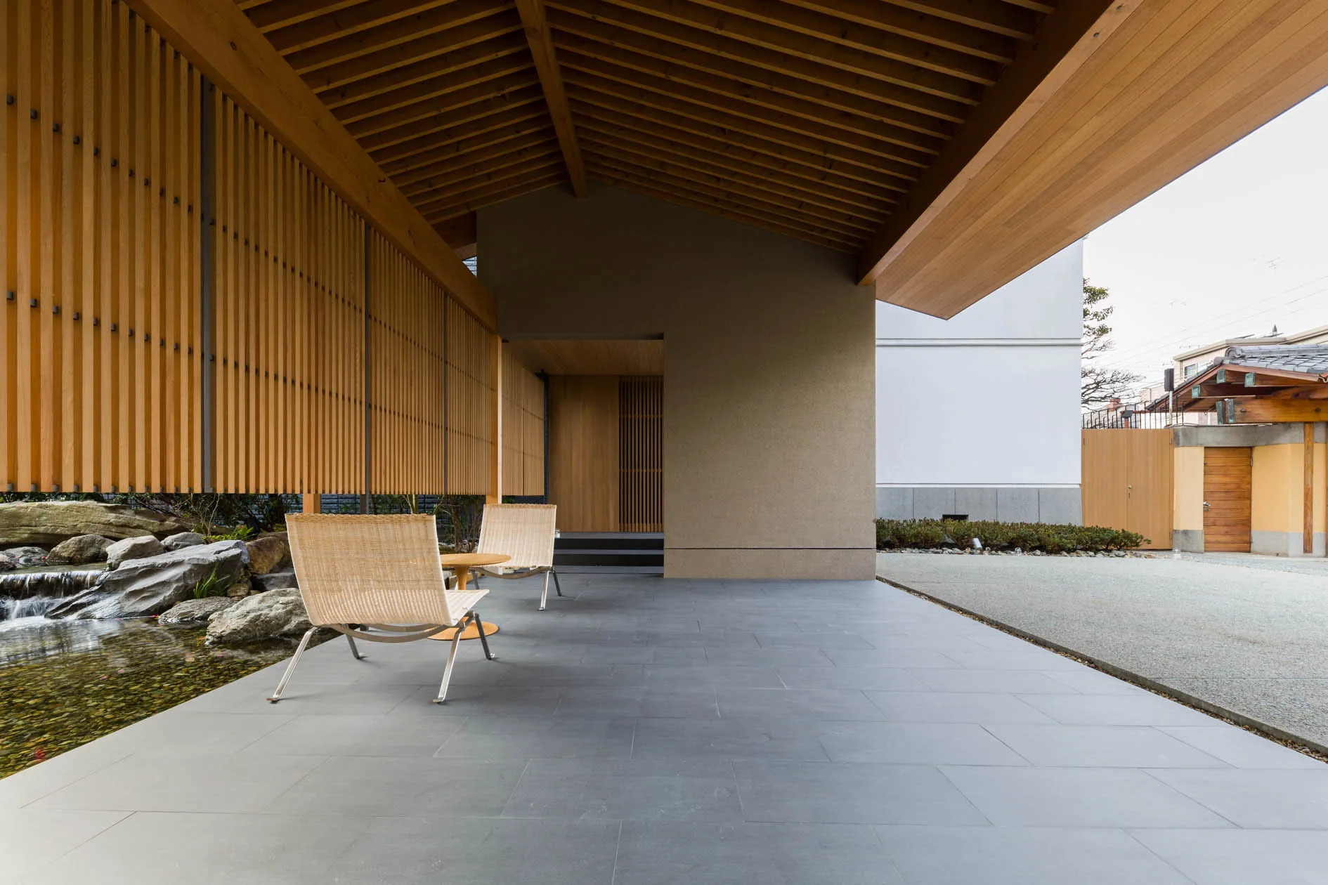 Two wicker chairs sit on a grey stone patio beneath an exposed wood slat ceiling next to a small garden water feature.