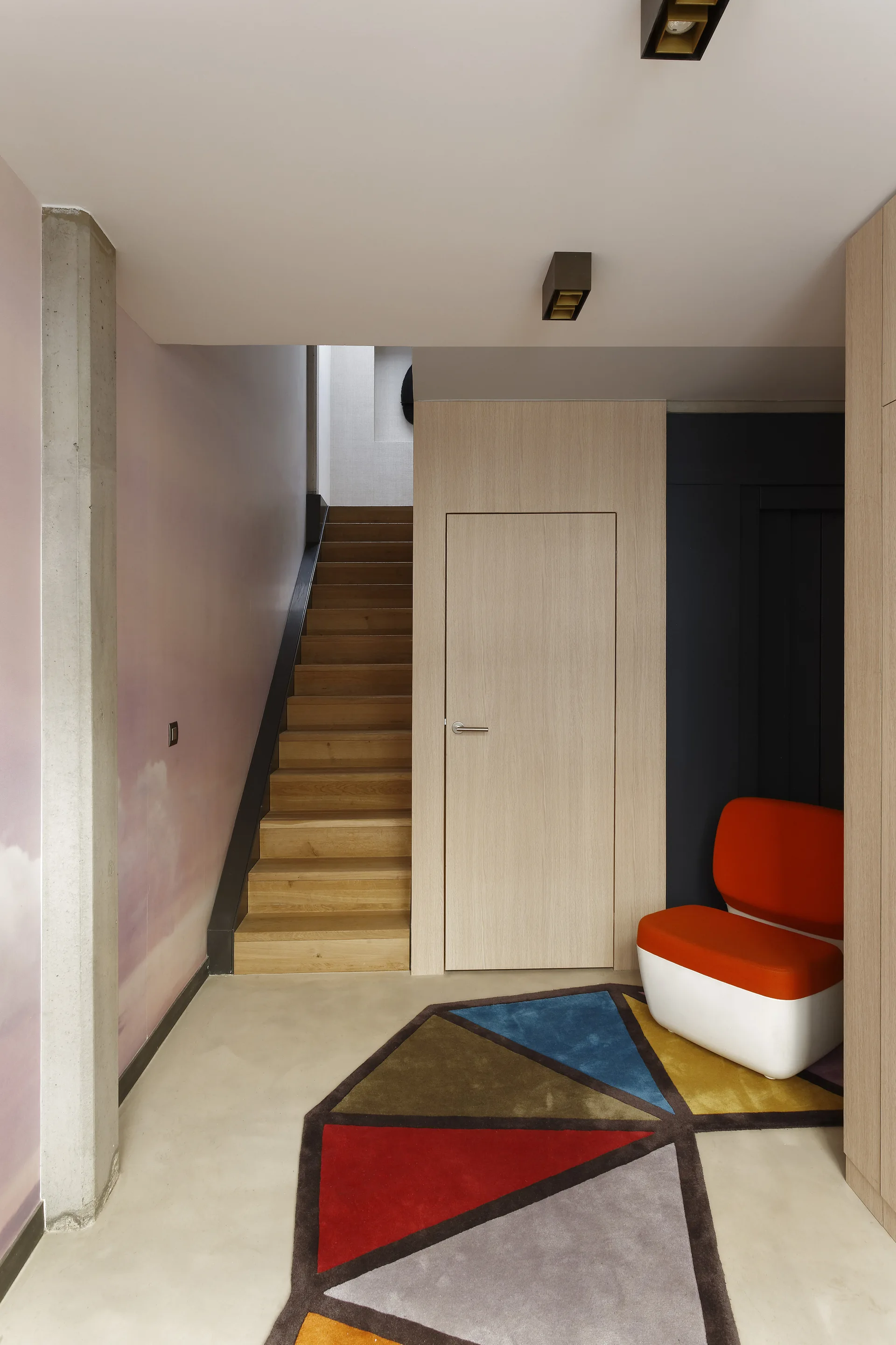 A modern entryway features a light wood staircase, a flush wood door, a colorful geometric rug, and an orange accent chair.