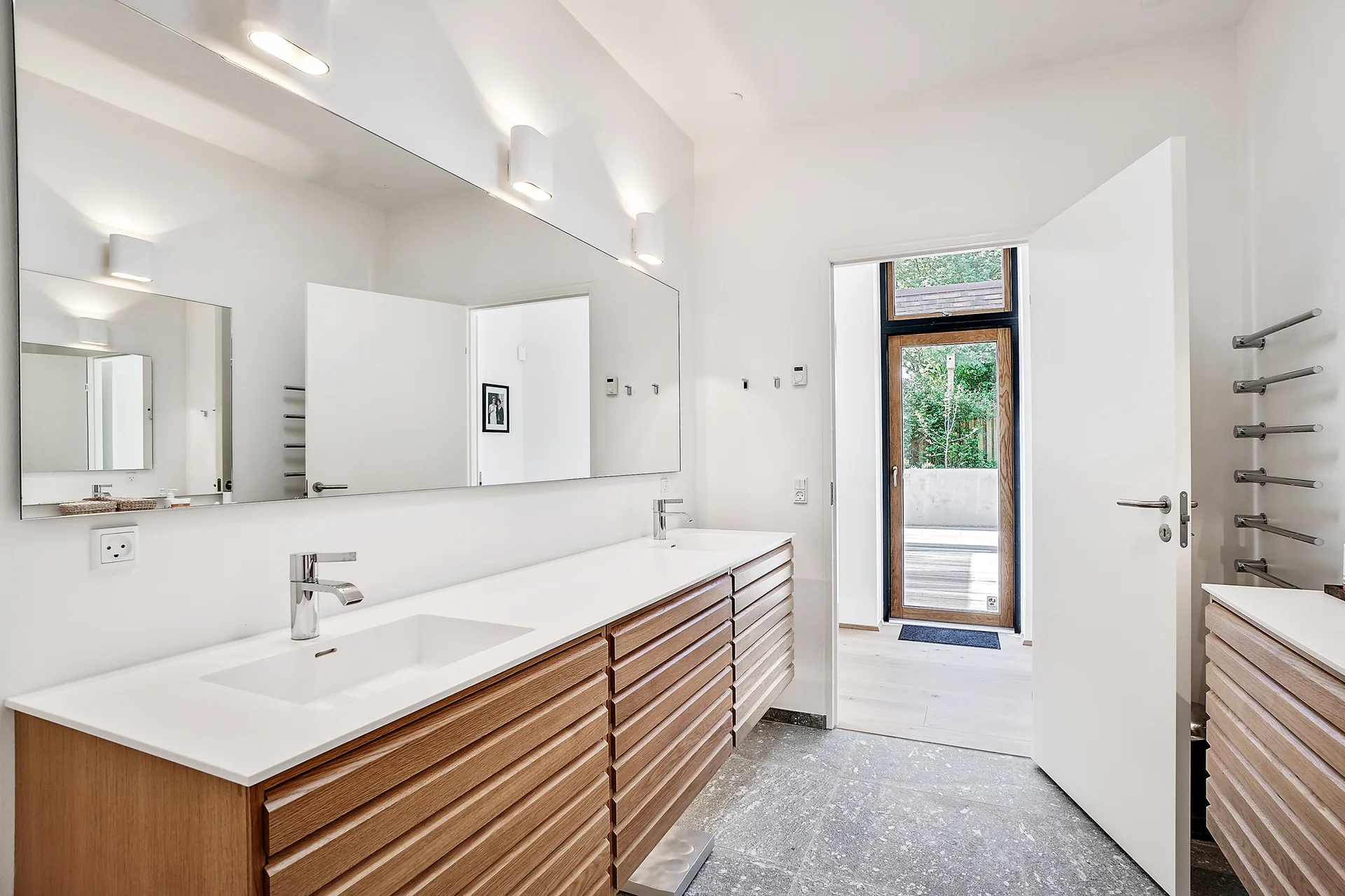 A modern bathroom with a light wood-slatted double vanity, a wide wall-mounted mirror, white countertops, and speckled grey stone tile floors.
