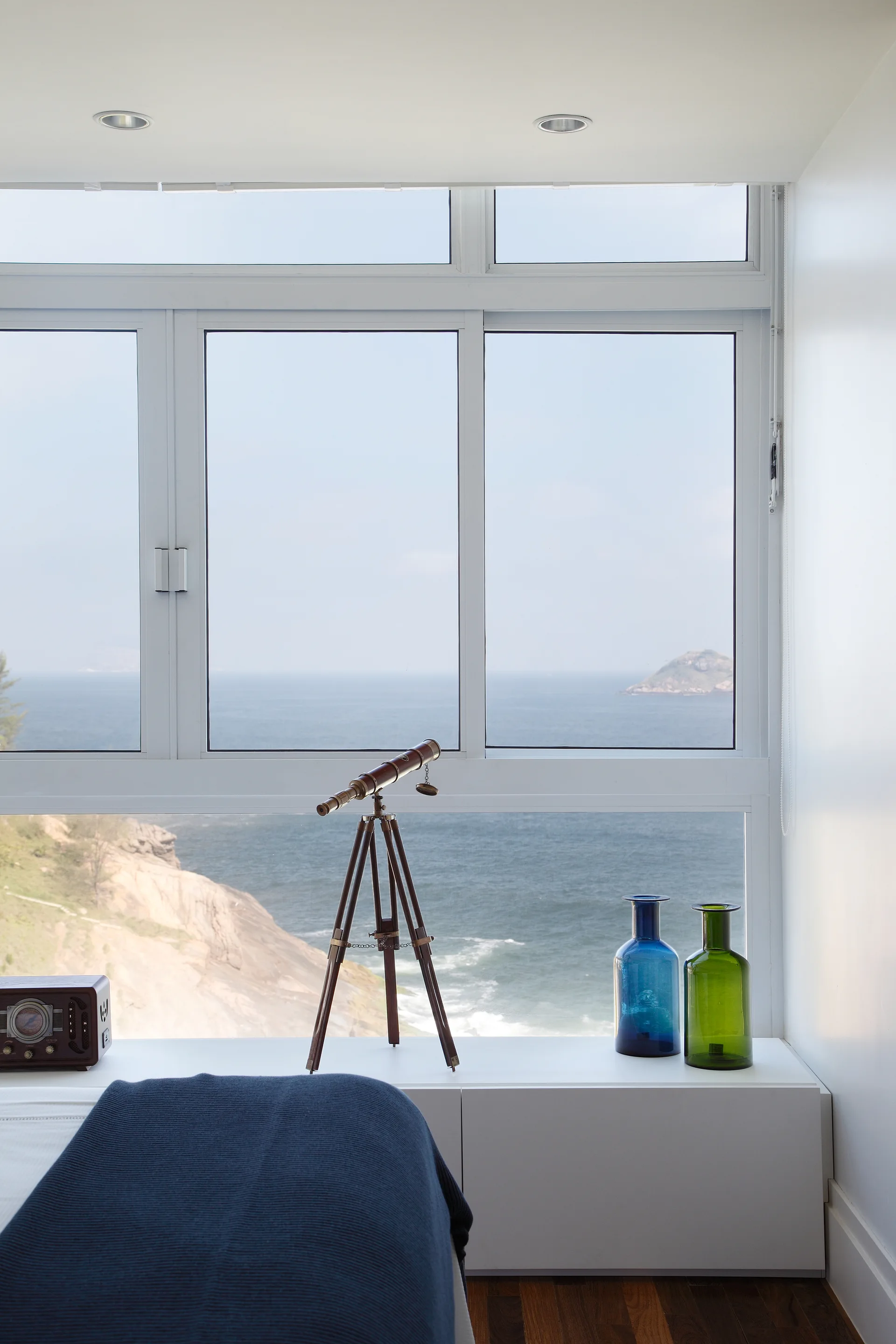 A minimalist bedroom window ledge displays a vintage telescope on a tripod and colorful glass vases against a scenic ocean backdrop.