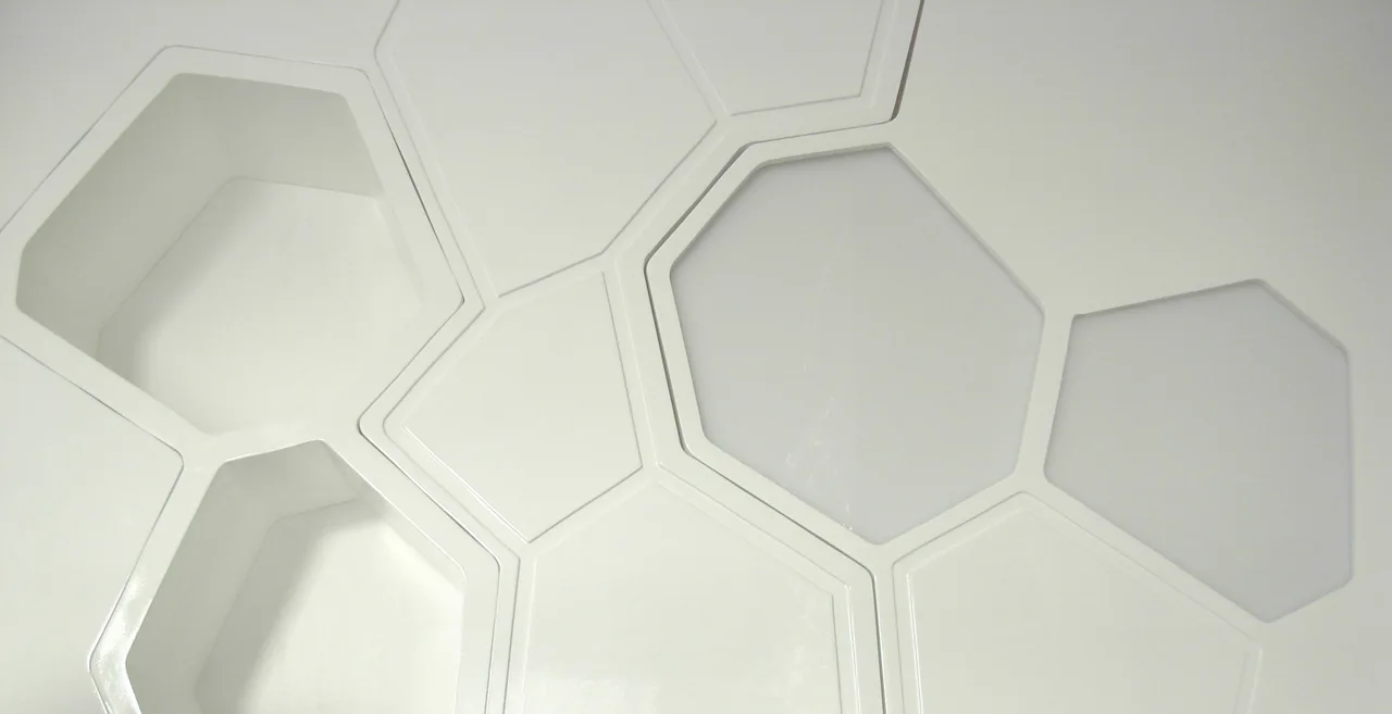 A close-up of a white modular wall panel system featuring a repeating geometric honeycomb pattern of recessed and flat hexagons.