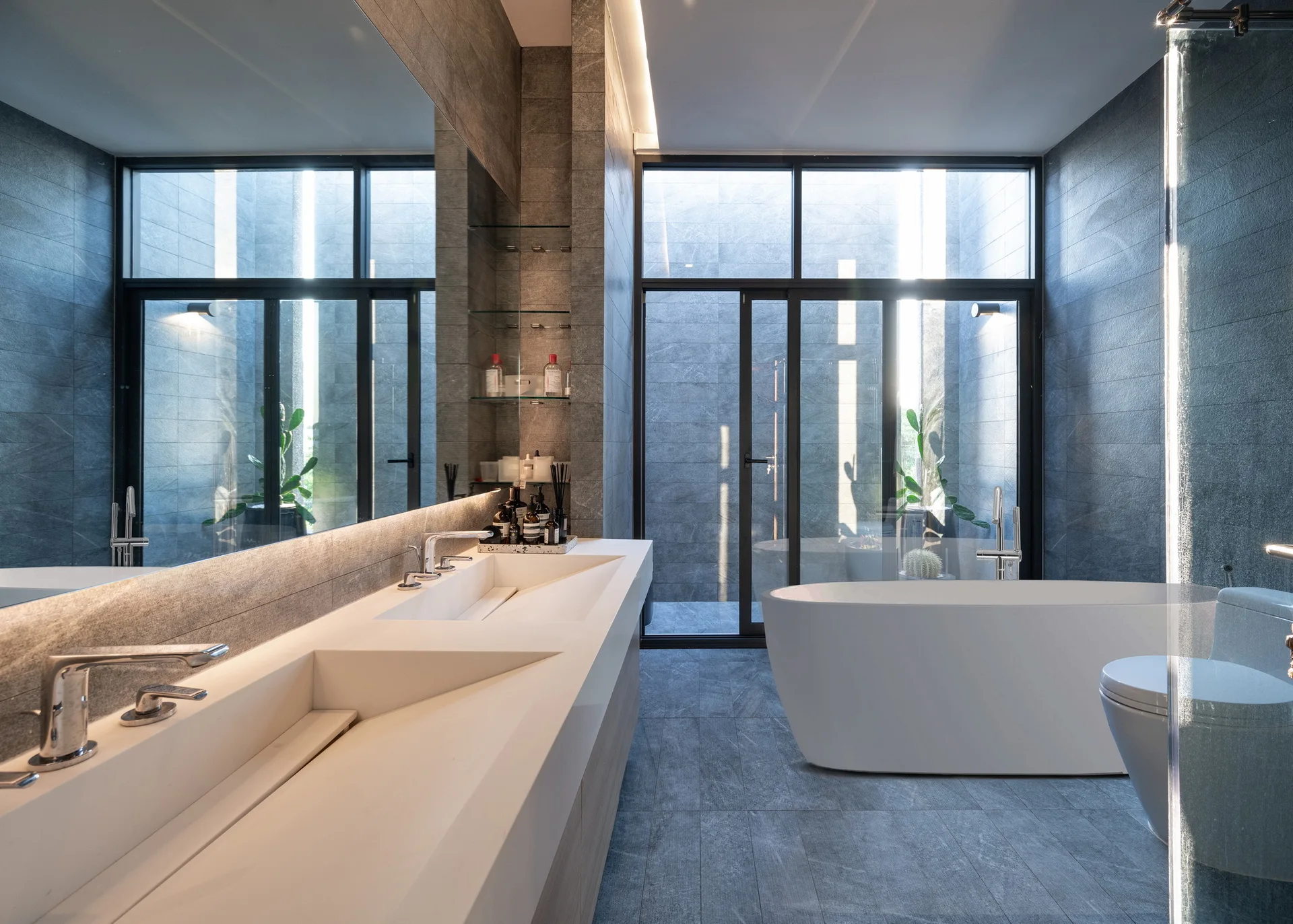 A modern grey stone bathroom featuring a long white double vanity with a backlit mirror, a freestanding bathtub, and floor-to-ceiling windows.