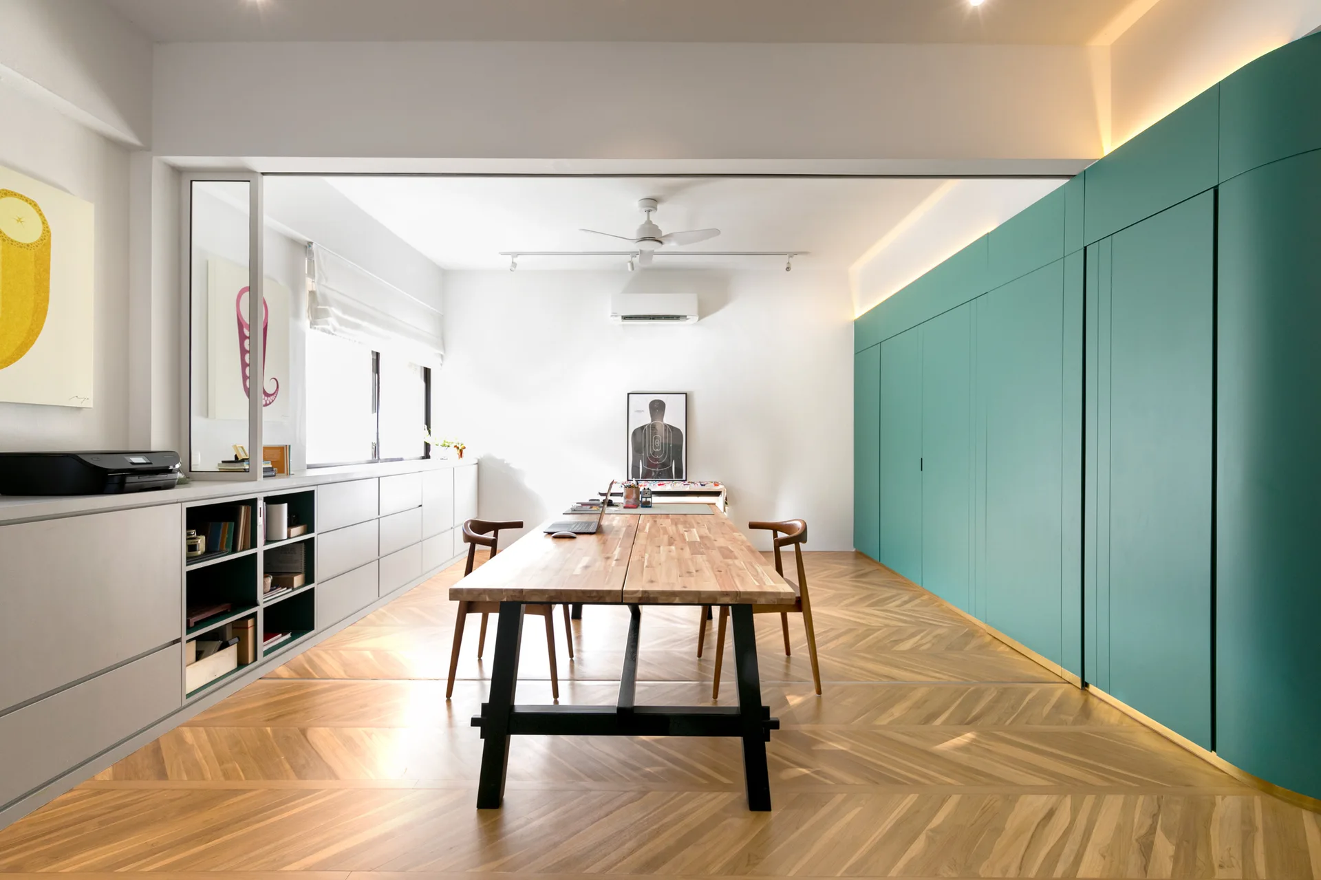 A spacious home office features a long wooden desk with black legs, herringbone wood floors, and a floor-to-ceiling teal storage wall.