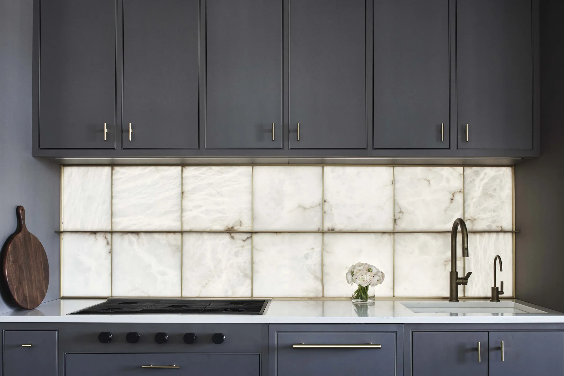 A modern kitchen features dark charcoal cabinets, a glowing backlit stone tile backsplash, a white countertop, and brass fixtures.