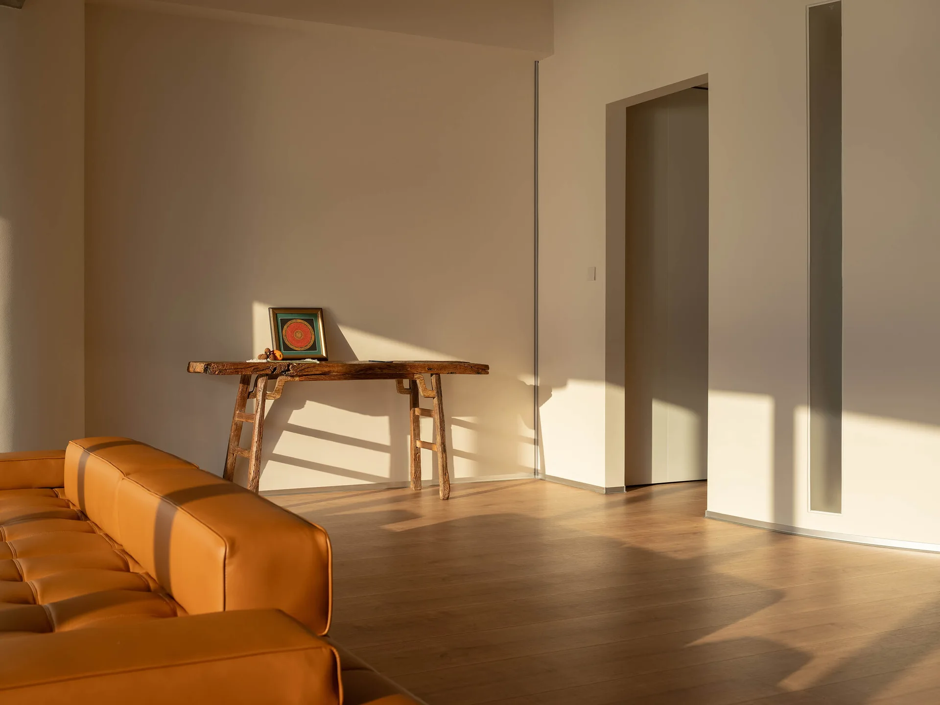 A minimalist living room featuring a tan leather sofa in the foreground and a rustic wooden console table against a sunlit beige wall.