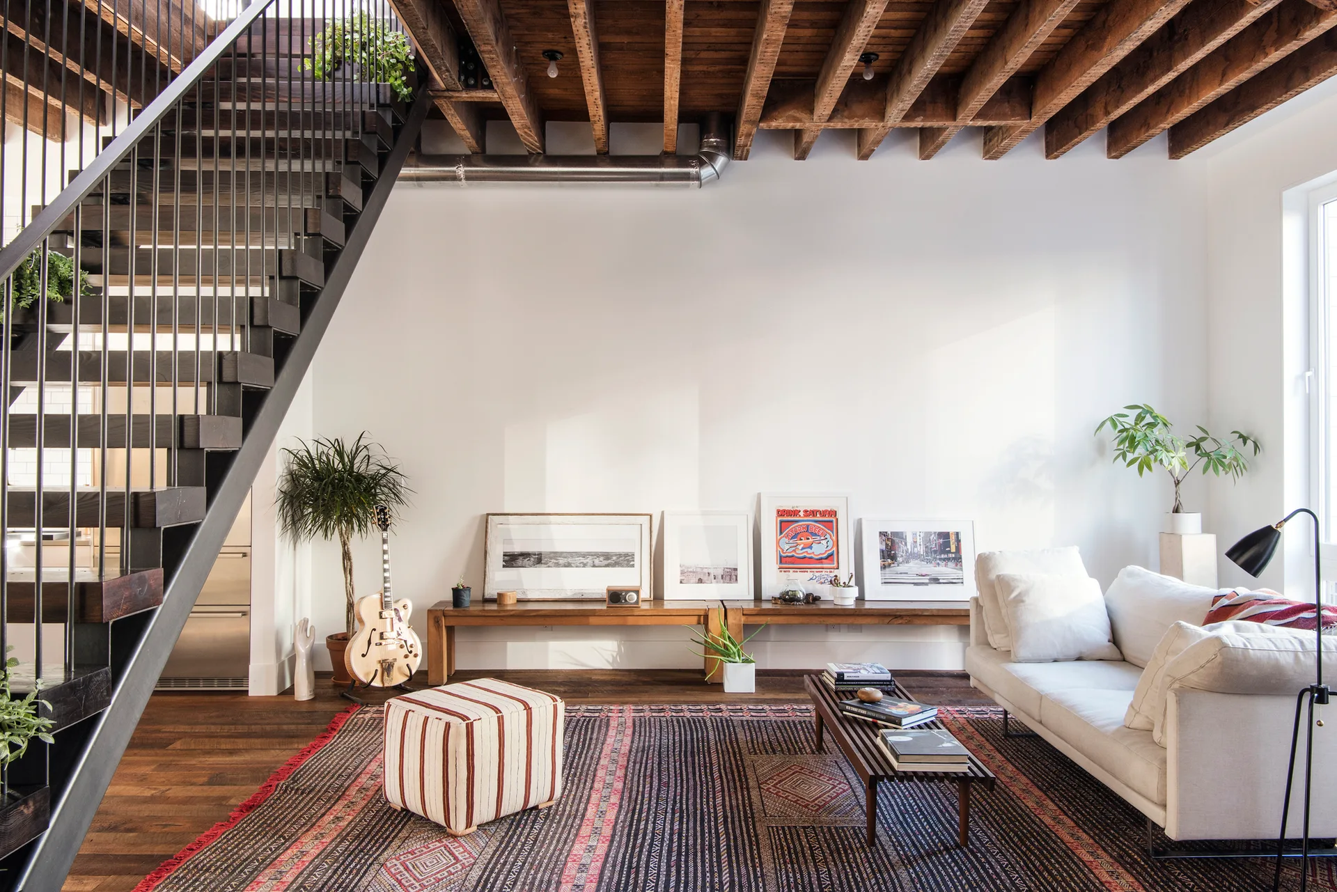 A living room features an open black metal staircase, a wood joist ceiling, a white sofa on a patterned rug, and art resting on a low wood bench.
