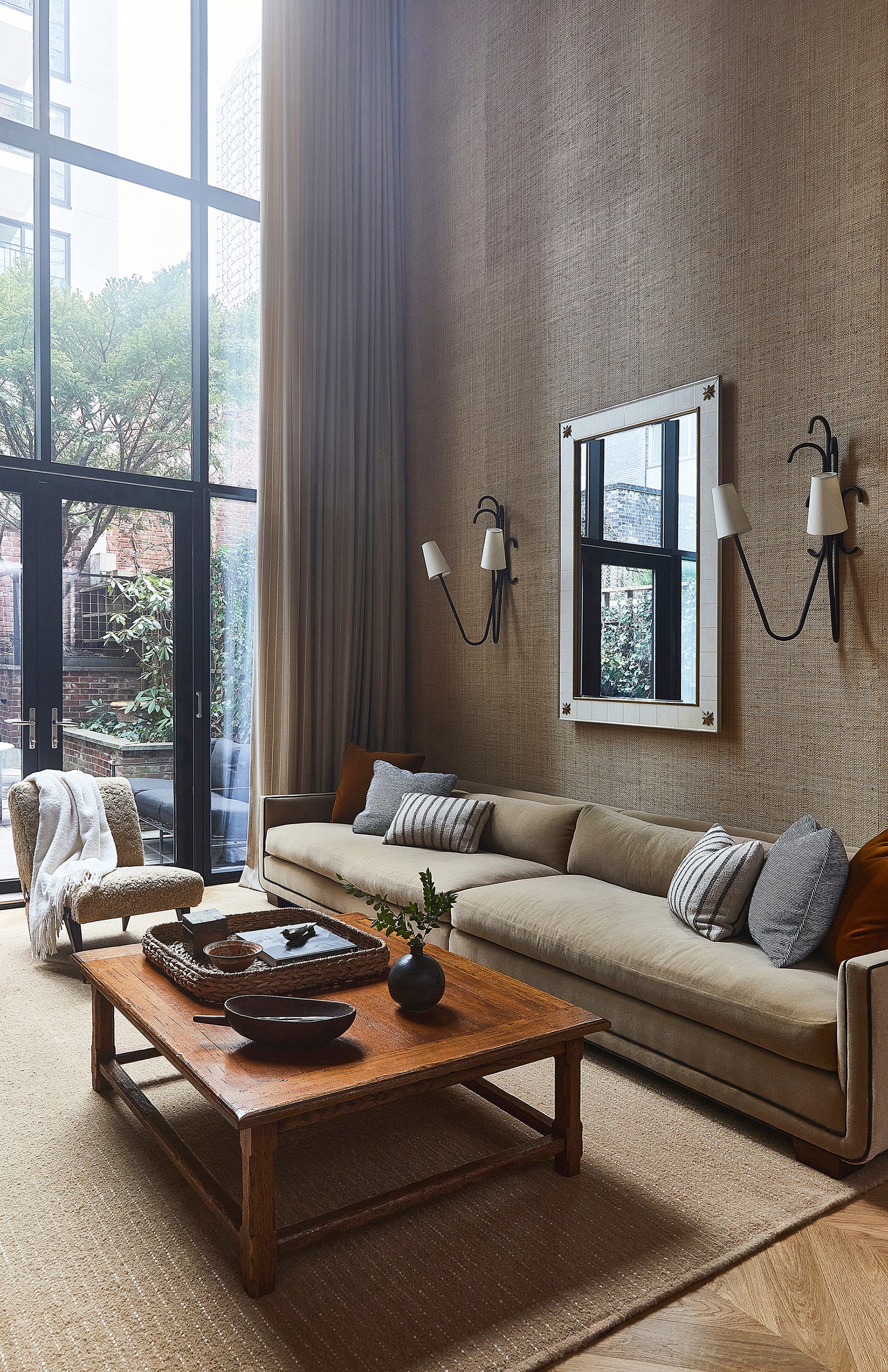 A high-ceilinged living room features a long beige sofa, a low wooden coffee table, a white wall mirror, and floor-to-ceiling windows.
