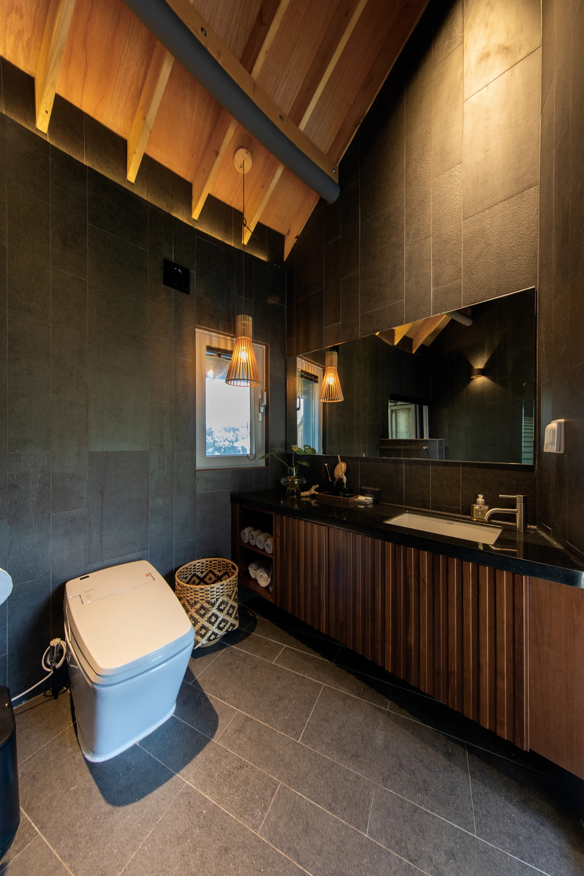 A modern bathroom with dark grey tiled walls, a wood-slatted vanity with a black countertop, and a sloped wood ceiling with exposed beams.