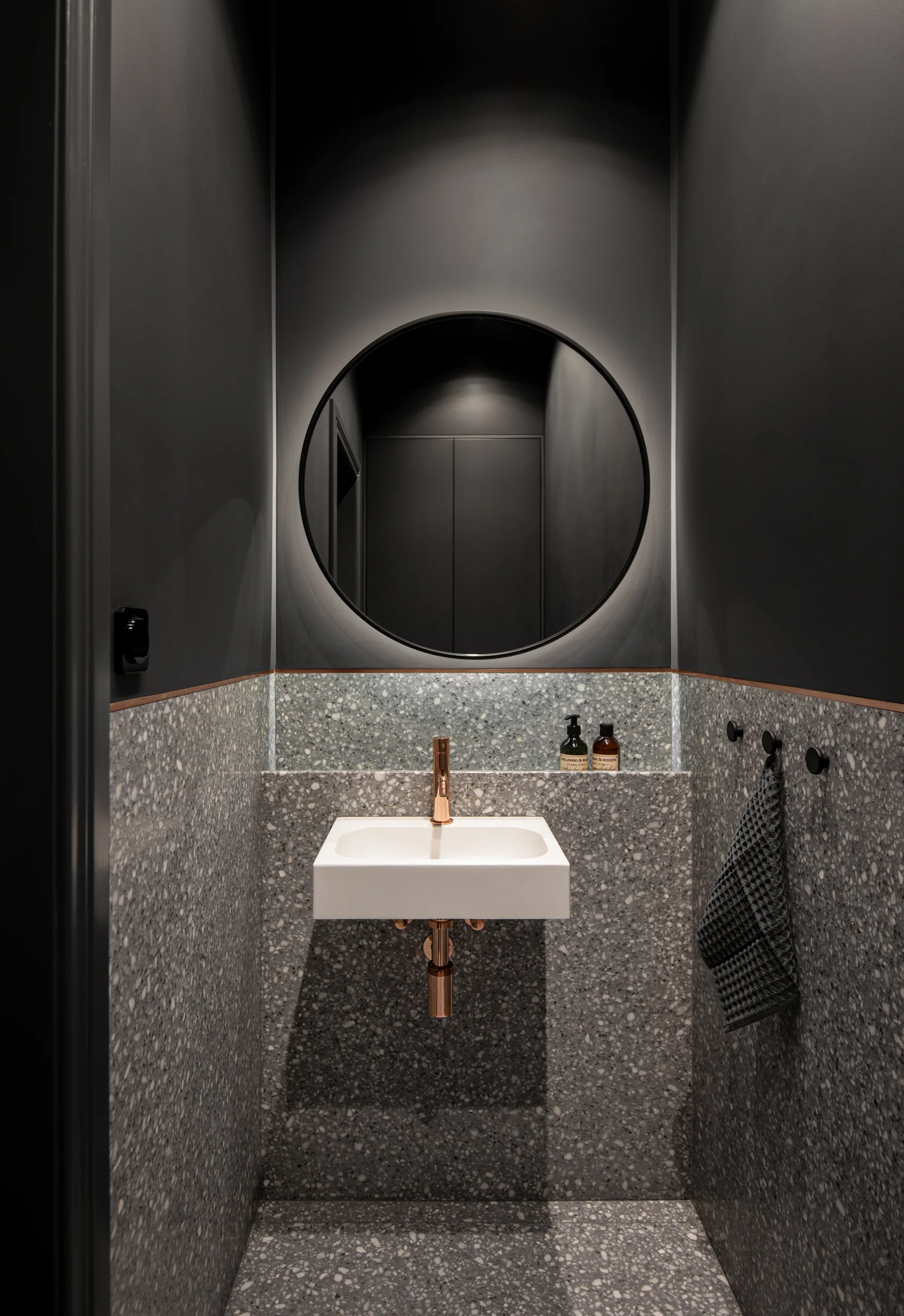 A small powder room featuring dark grey painted walls, a round backlit mirror, a white wall-mounted basin with copper fixtures, and grey terrazzo flooring.