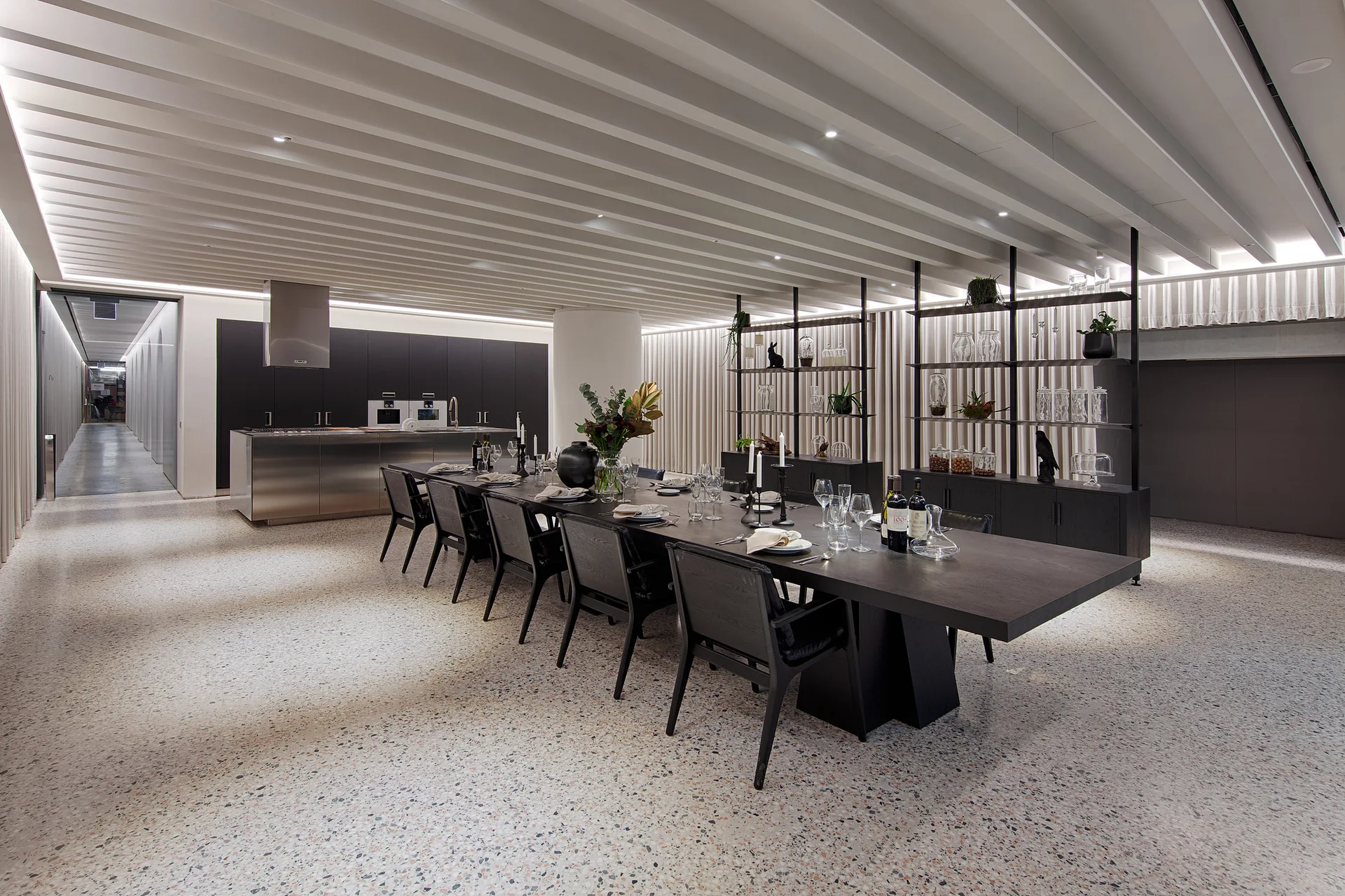 A long black dining table sits on speckled terrazzo flooring beneath a white slatted ceiling in a modern open kitchen and dining area.