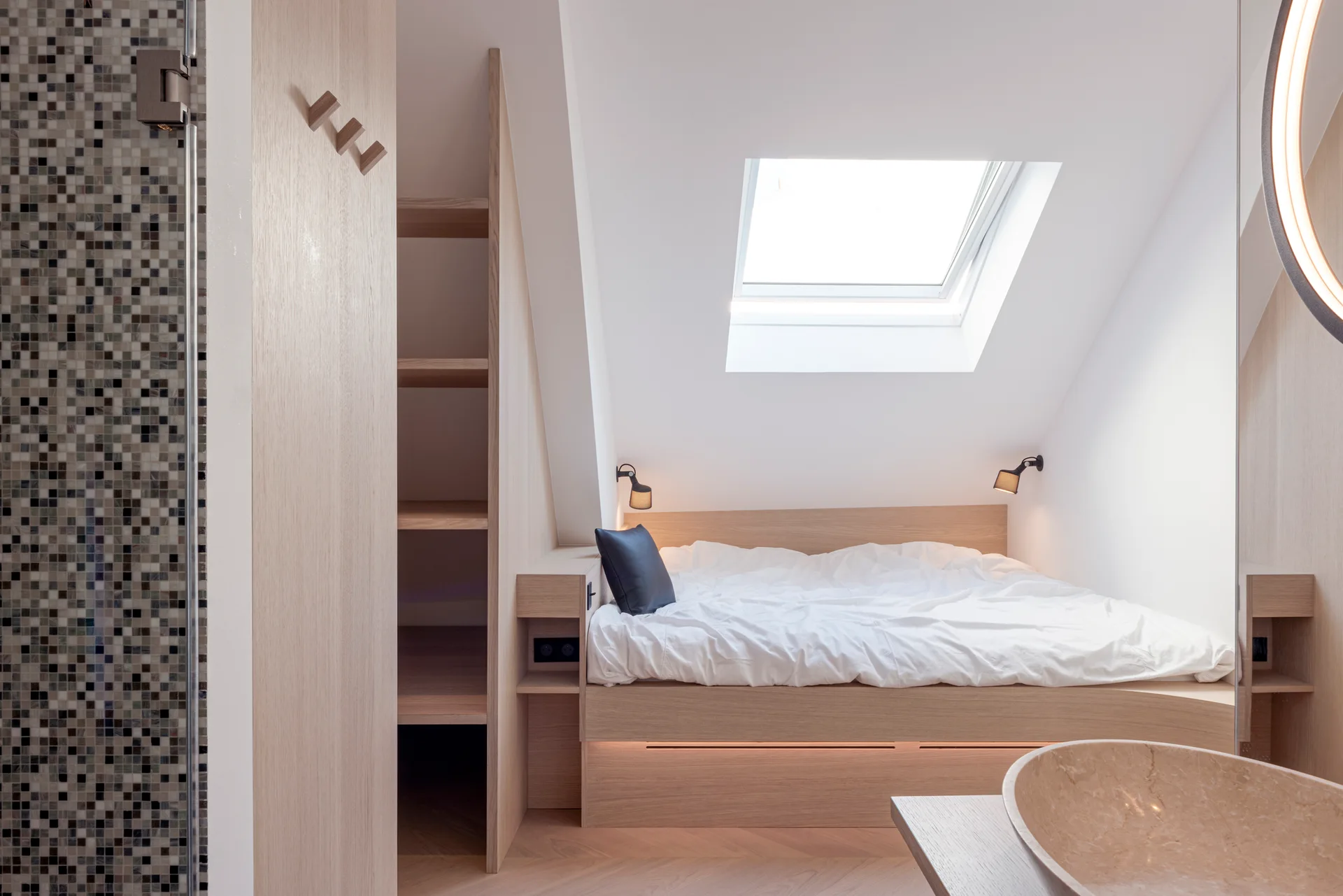A modern attic bedroom featuring a built-in light wood platform bed beneath a sloped ceiling with a skylight and custom open shelving.
