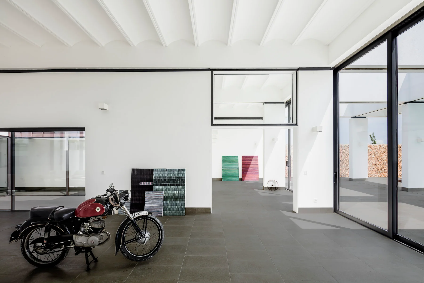 A spacious minimalist room features white walls, dark floor tiles, ribbed ceiling beams, and a vintage red and black motorcycle parked indoors.