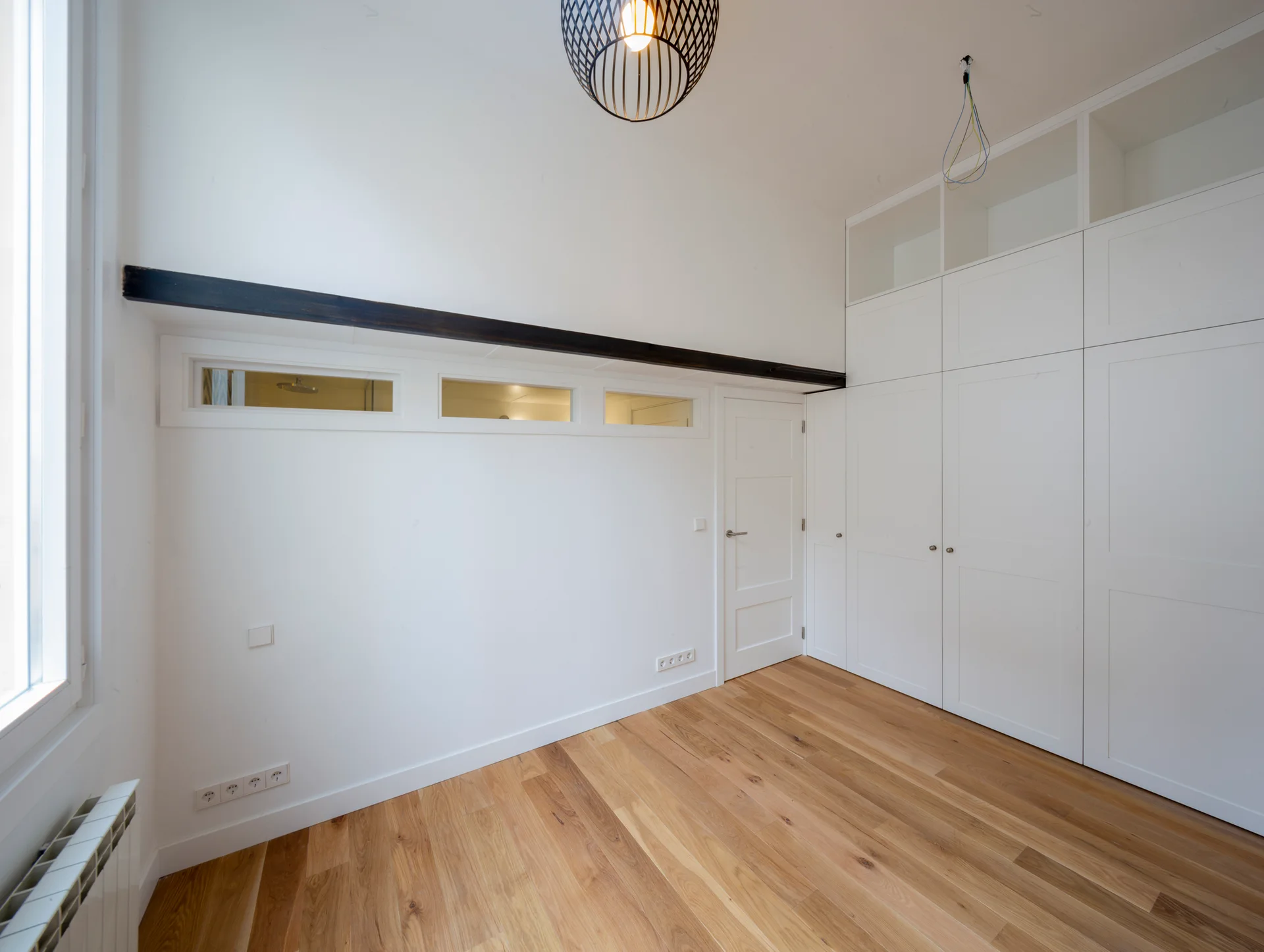 An empty bedroom features light wood floors, floor-to-ceiling white built-in closets, an interior door, and small transom windows beneath a dark beam.