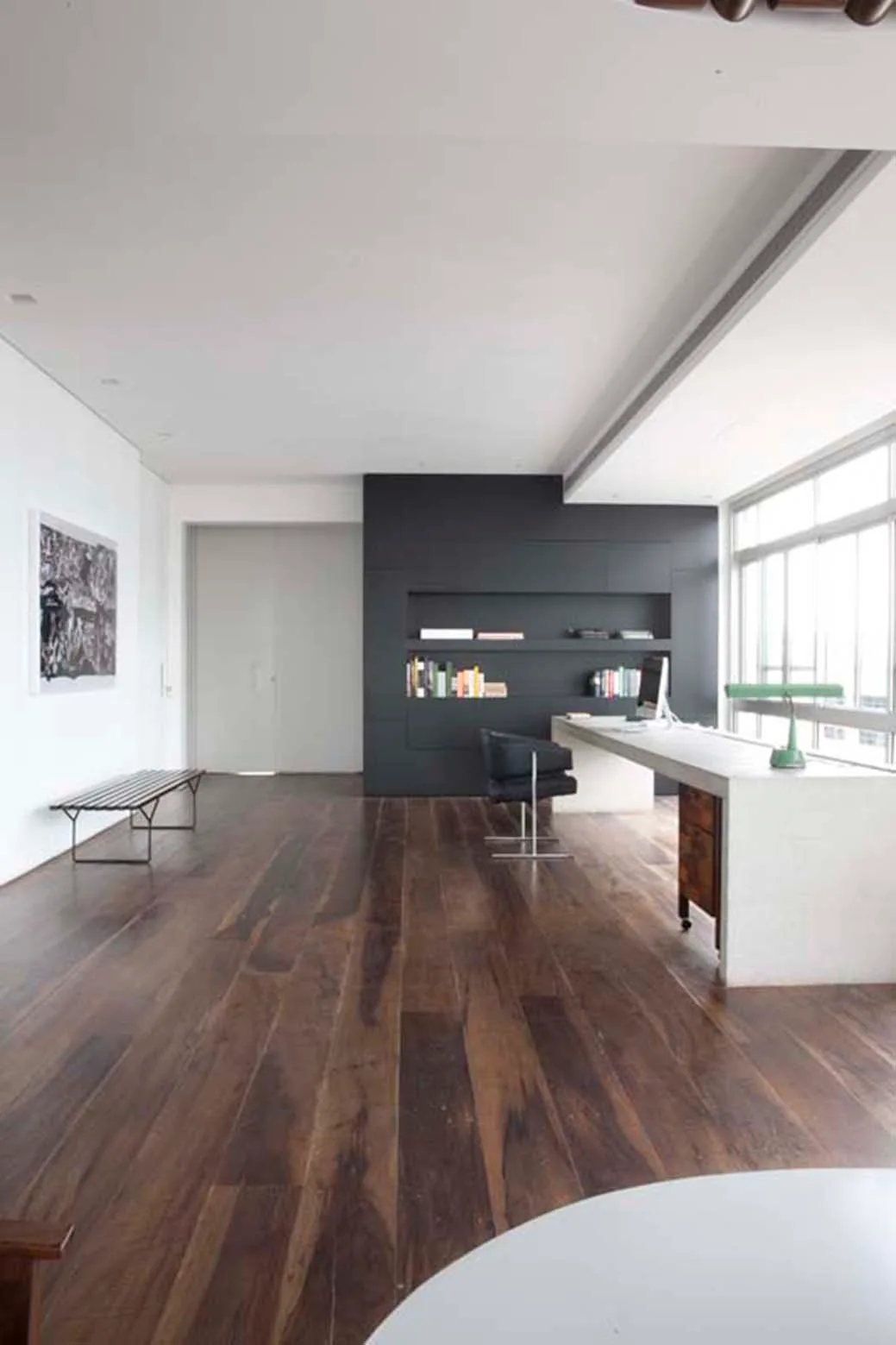 A minimalist home office featuring dark wide-plank wood floors, a long white desk, and a dark accent wall with recessed built-in shelves.