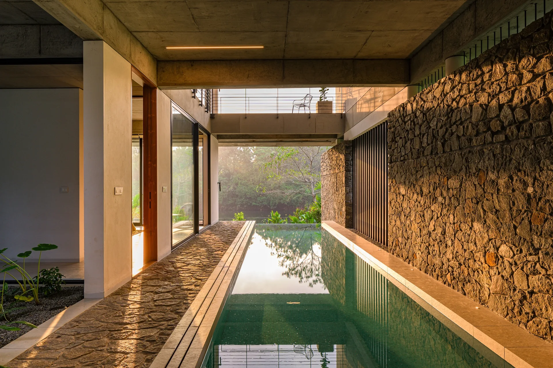 A narrow lap pool sits within a concrete structure, flanked by a rough stone wall and glass doors, looking out toward a river landscape.