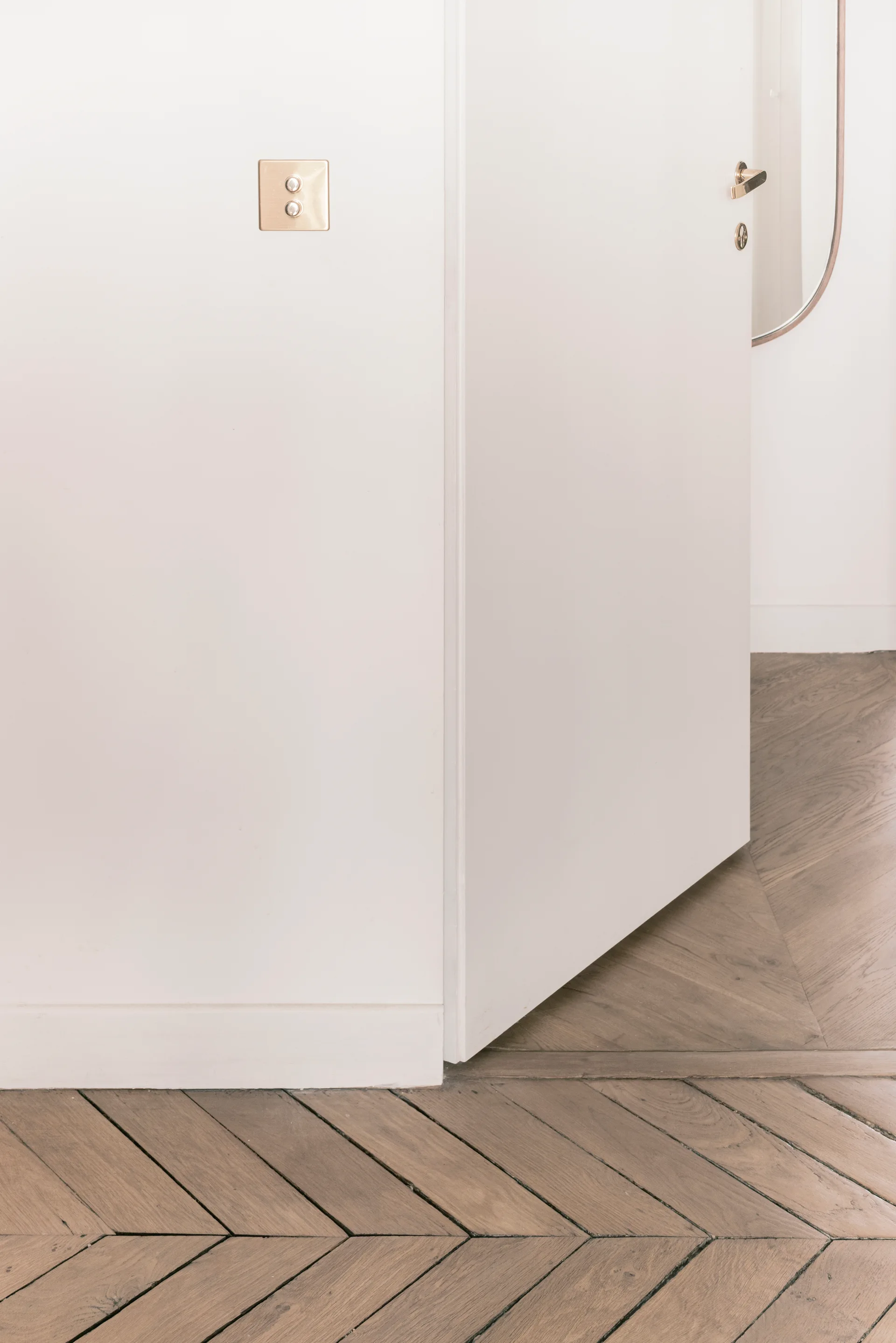A partially open white door and wall corner with brass hardware set above a classic chevron-patterned wood floor.