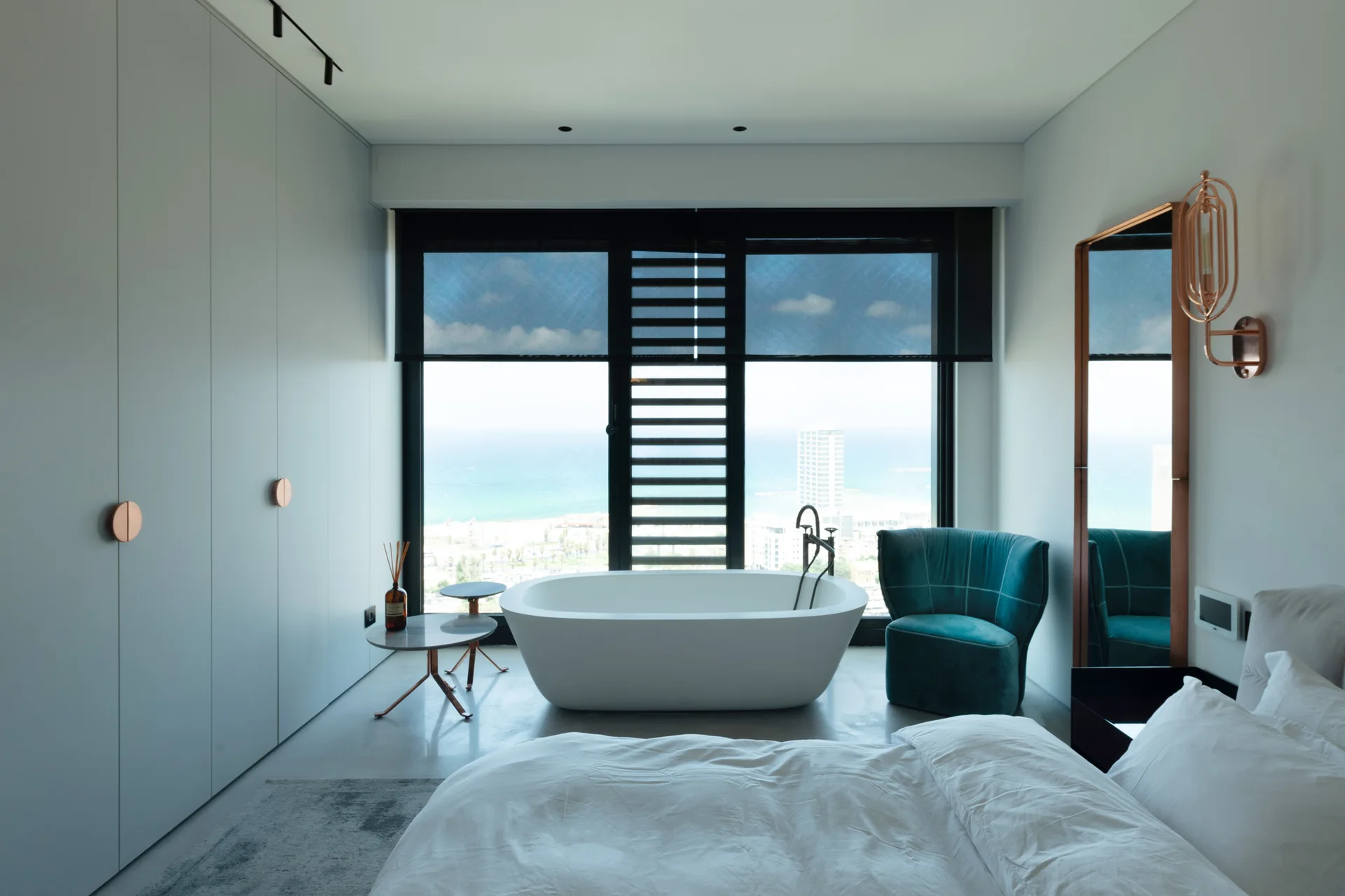 A minimalist grey bedroom features a white freestanding bathtub positioned before a large window overlooking the ocean, with a bed in the foreground.