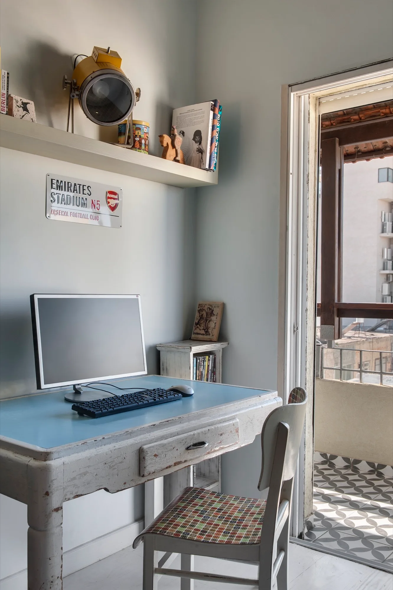 A distressed wooden desk with a computer monitor stands in a small home office next to a glass balcony door showing geometric floor tiles.