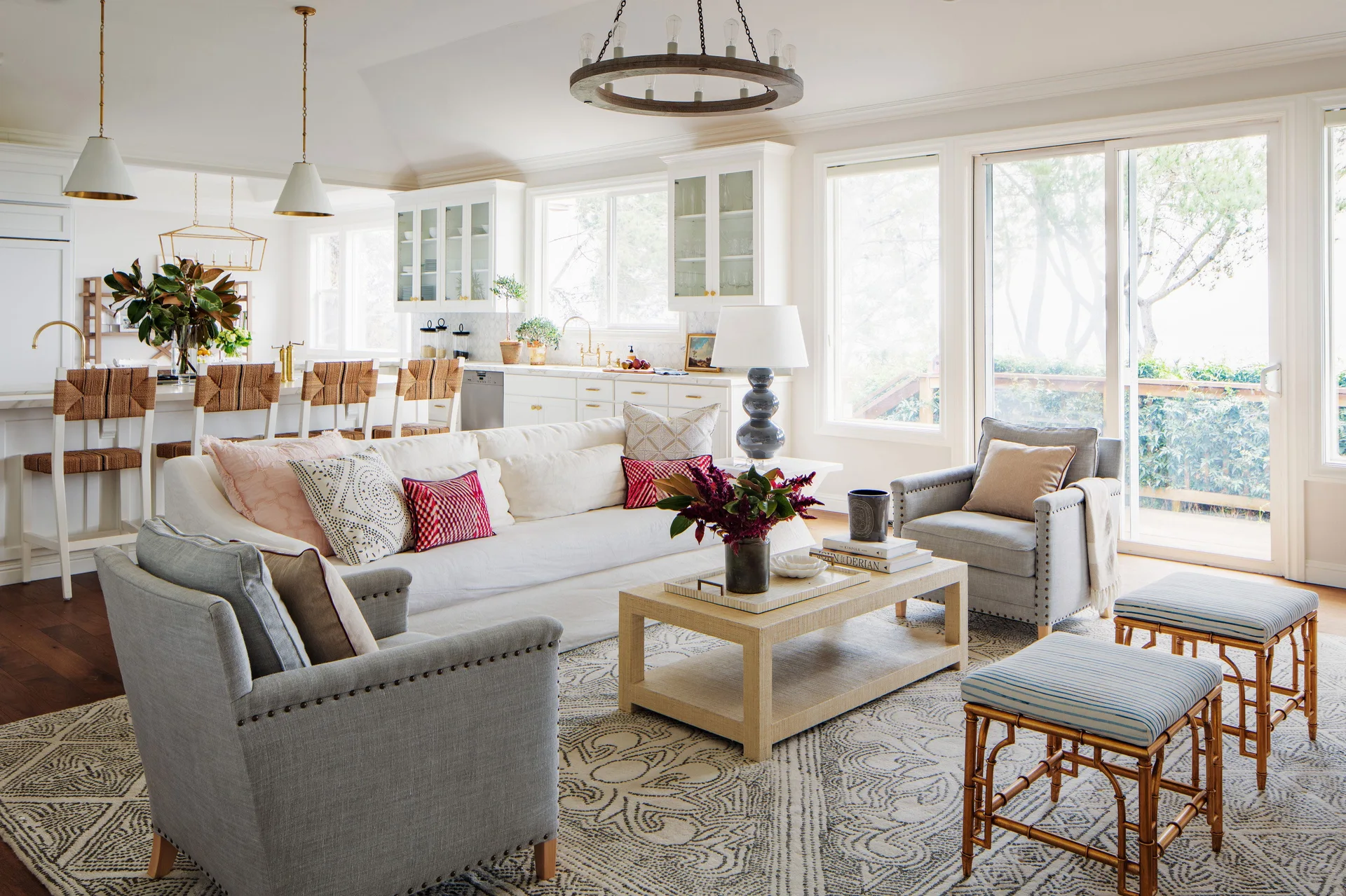 A bright transitional living room featuring a white sofa, two gray nailhead trim armchairs, and a large patterned rug, opening to a white kitchen.