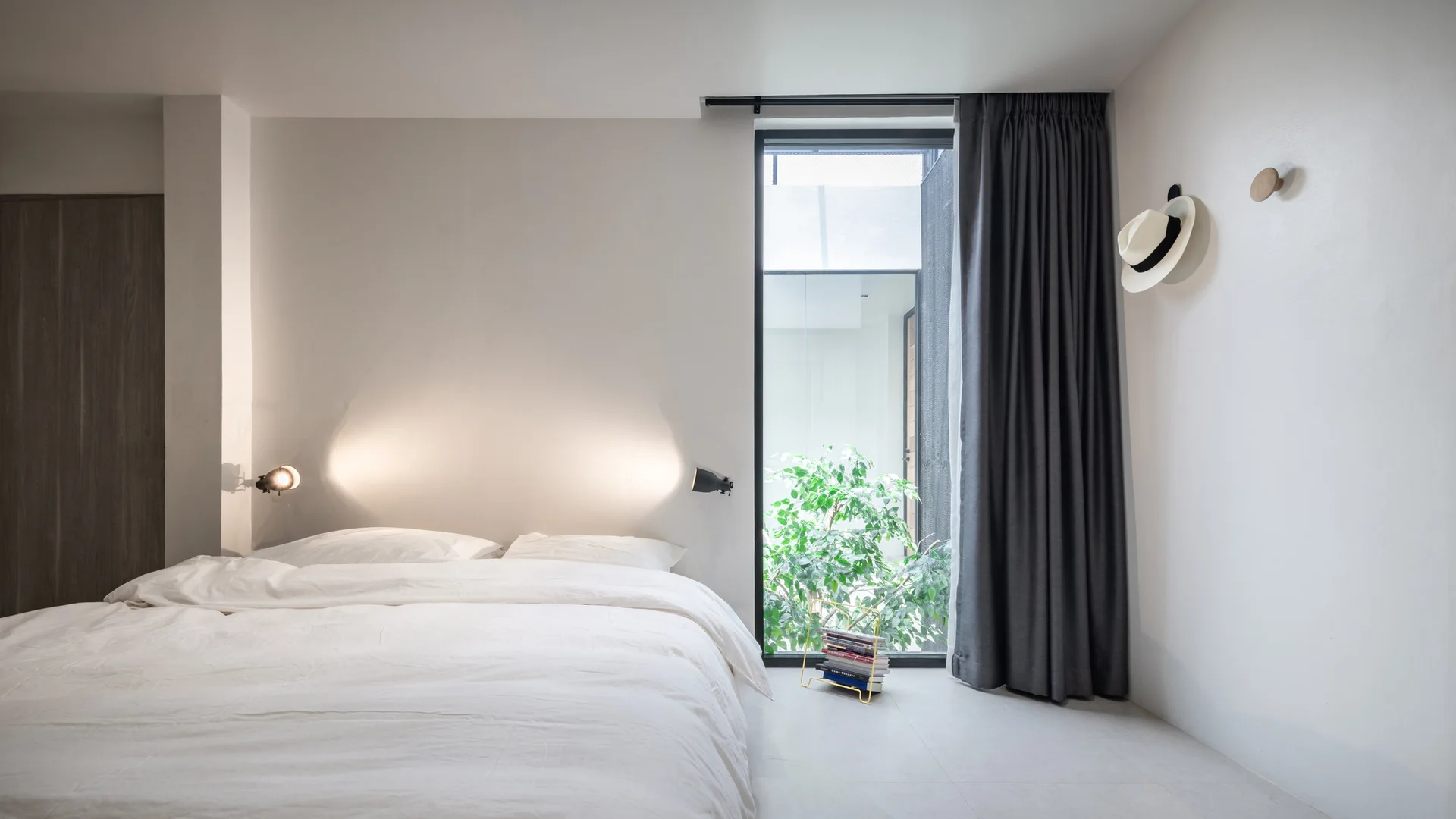 A minimalist white bedroom with a low bed, a narrow floor-to-ceiling window showcasing green foliage, dark grey drapes, and illuminated wall sconces.