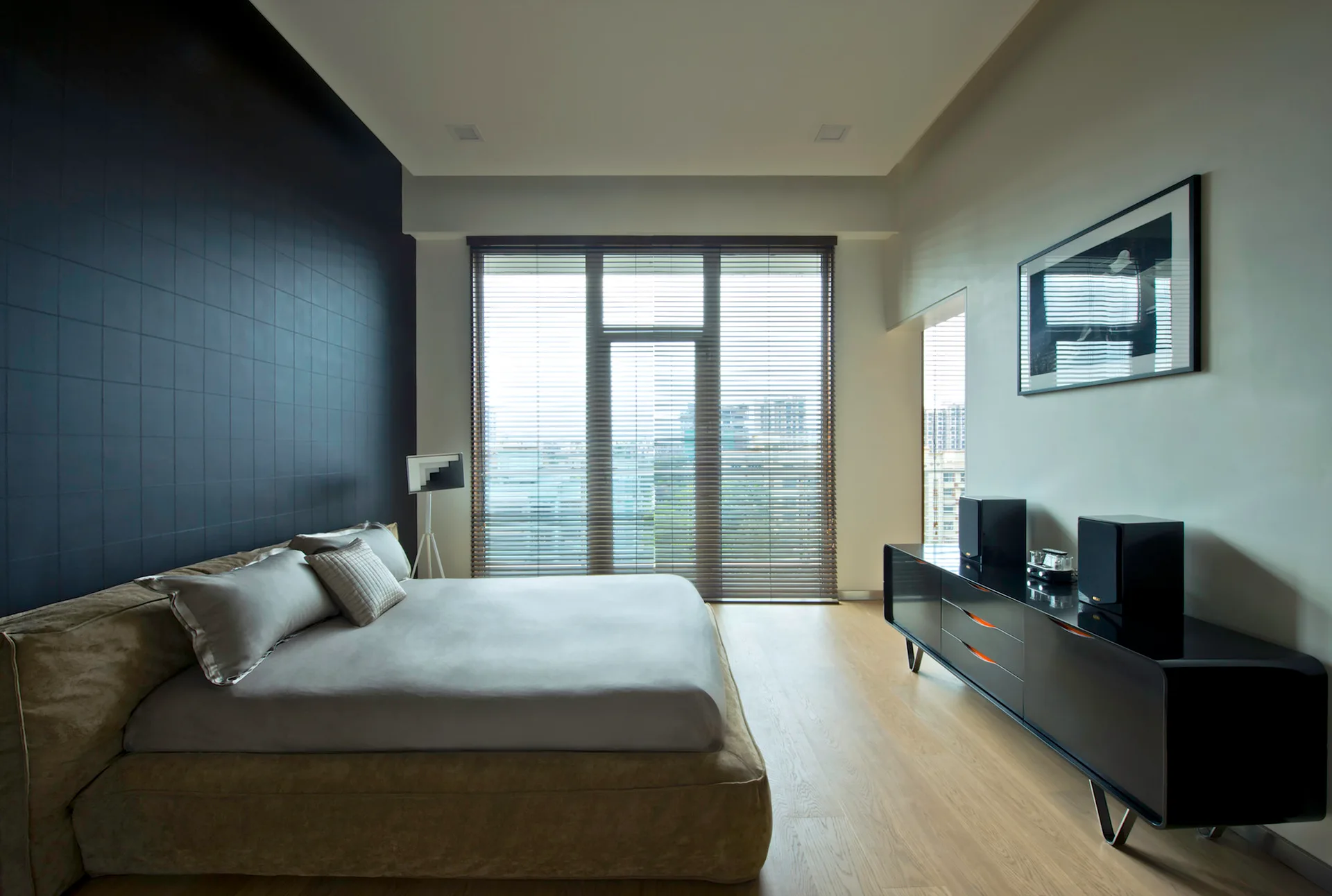 A contemporary bedroom featuring a low upholstered bed set against a dark tiled wall opposite a glossy black dresser next to windows with blinds.