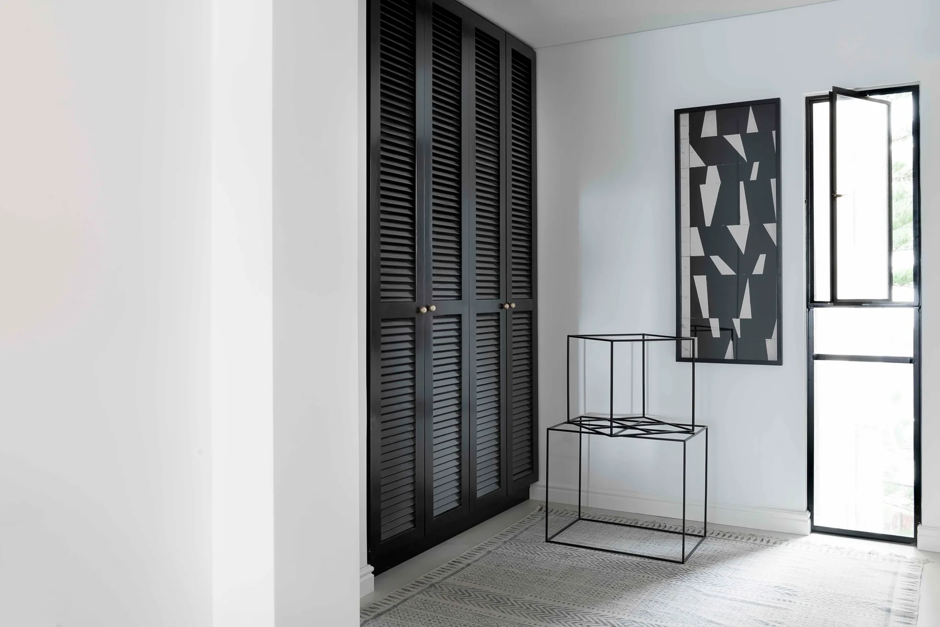 A minimalist room corner features tall black louvered closet doors, a metal wire cube sculpture, geometric wall art, and a patterned rug.