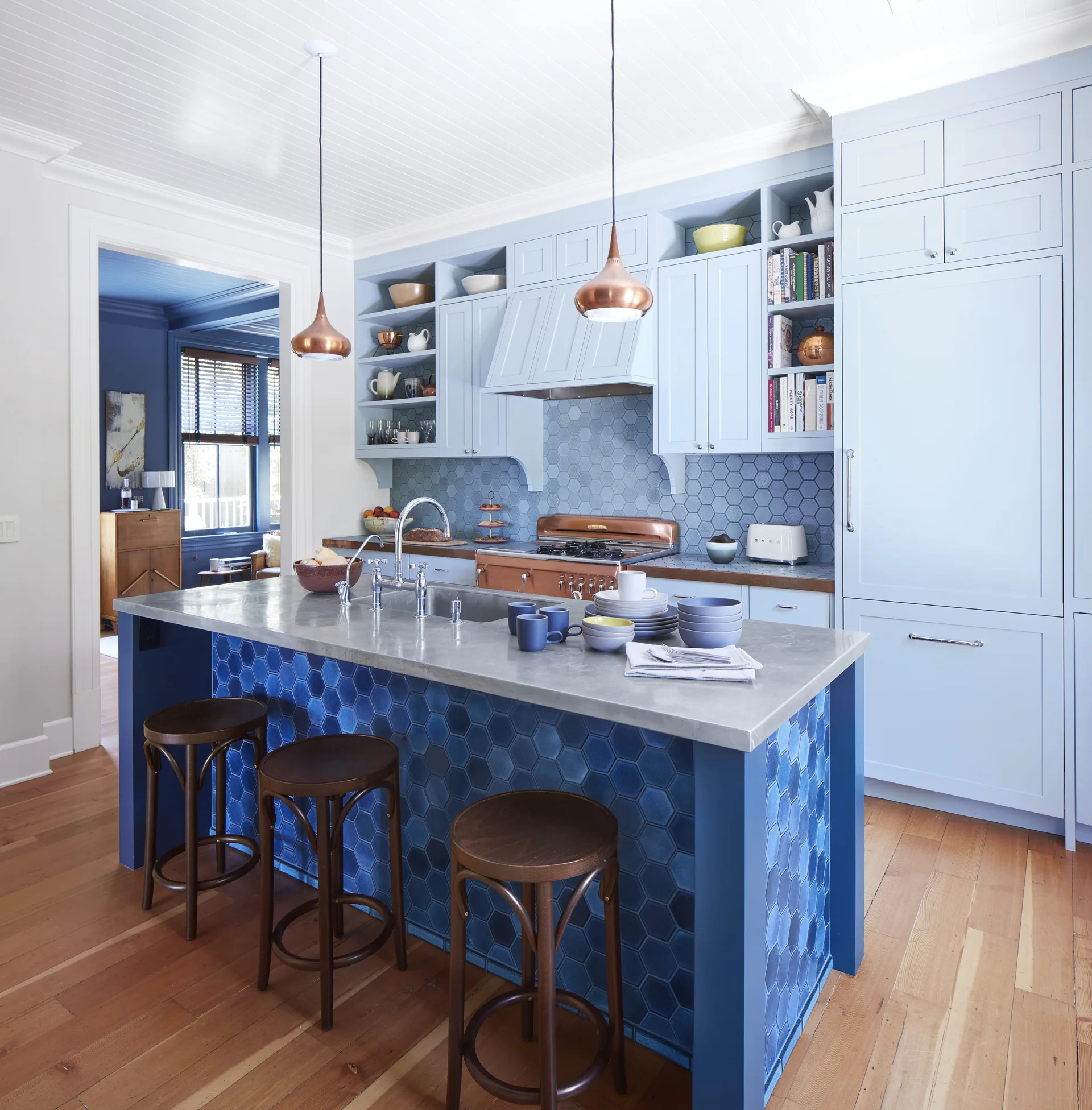 A light blue kitchen features a central island faced in blue hexagonal tiles, three dark wood stools, and copper pendant lights.