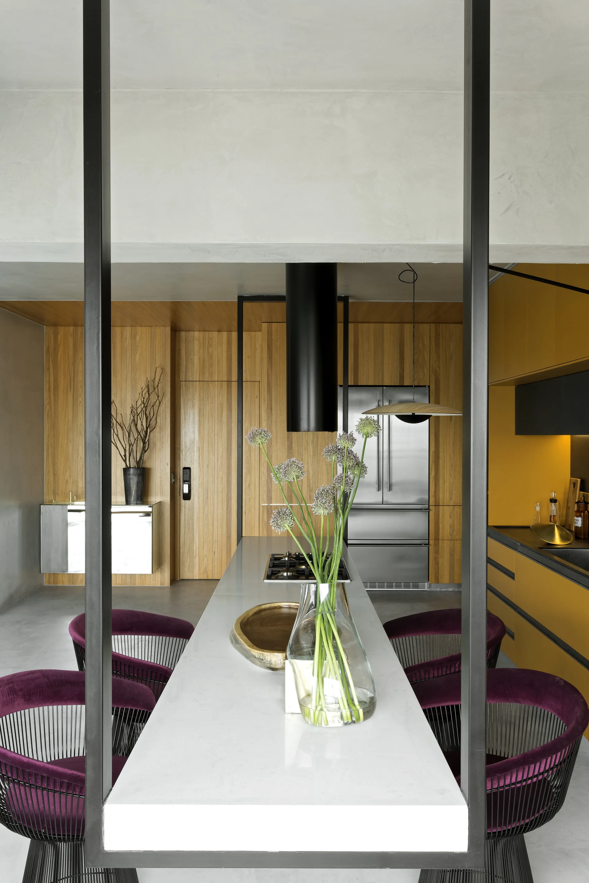 A long stone kitchen island with plum velvet chairs is framed by black metal columns in a kitchen with wood paneling and yellow cabinets.