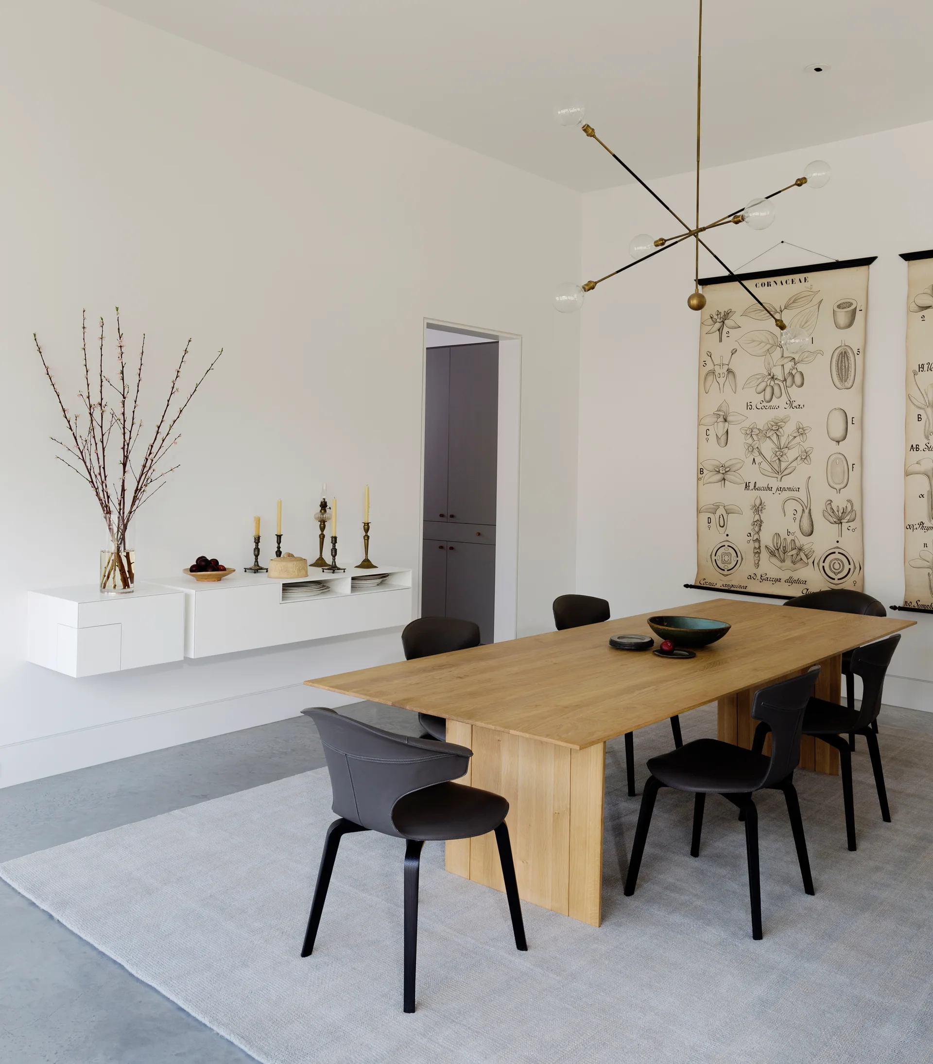 A minimalist dining room features a light wood table, dark grey chairs, a white floating console, and vintage botanical scrolls on white walls.