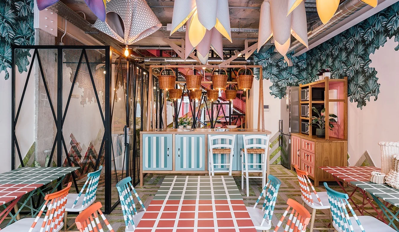 A playful dining space features colorful grid-patterned tables, spindle-back chairs, a striped kitchen counter, and sculptural petal-shaped pendant lights.