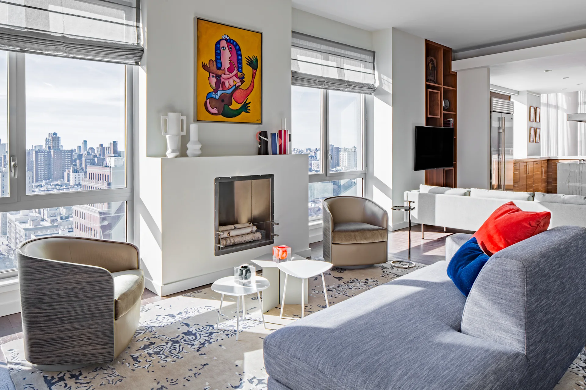 A bright living room featuring a grey sectional, two curved armchairs, a white fireplace with a yellow painting, and large windows with city views.