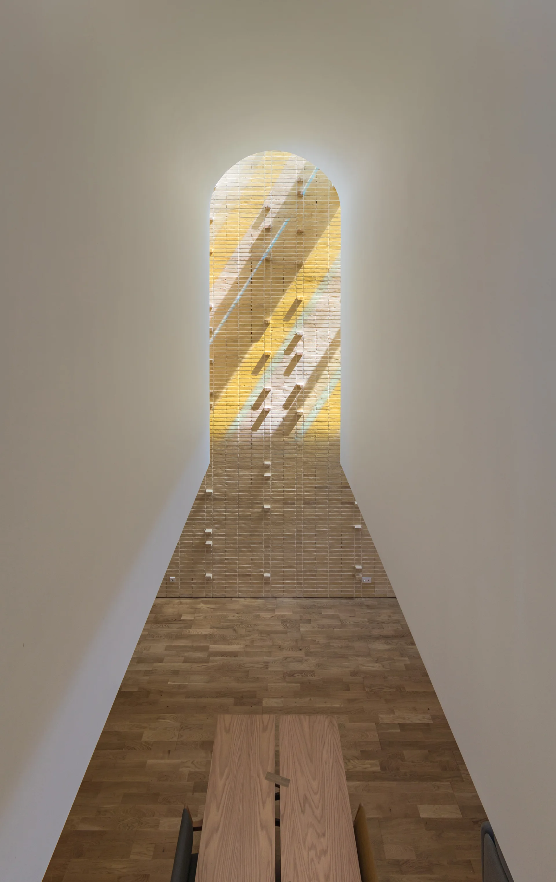 A long room features white walls, a parquet wood floor, a wooden table, and an arched brick accent wall with protruding bricks and yellow stripes.